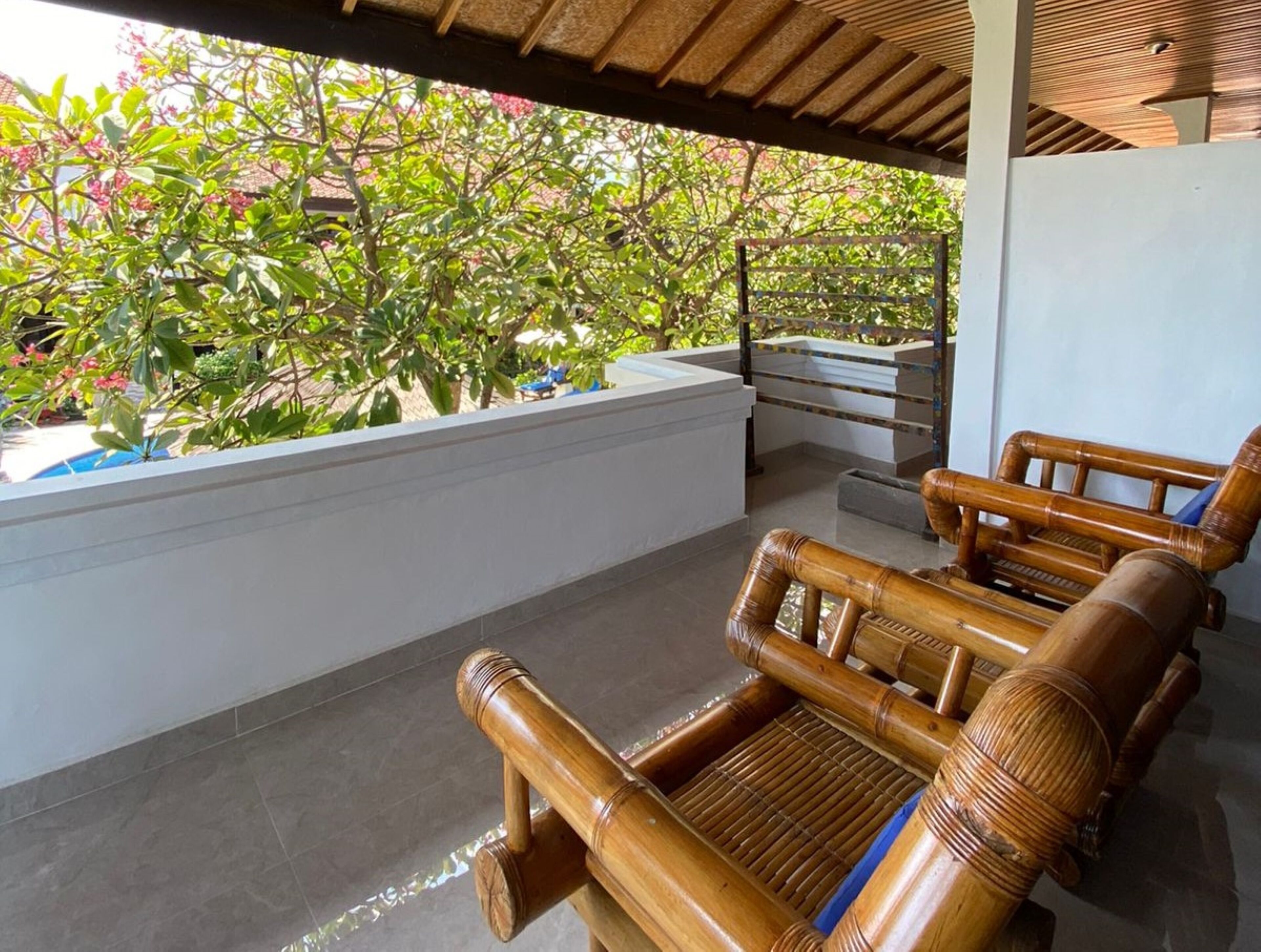 A covered outdoor seating area with bamboo furniture, a white wall, and a view of greenery.