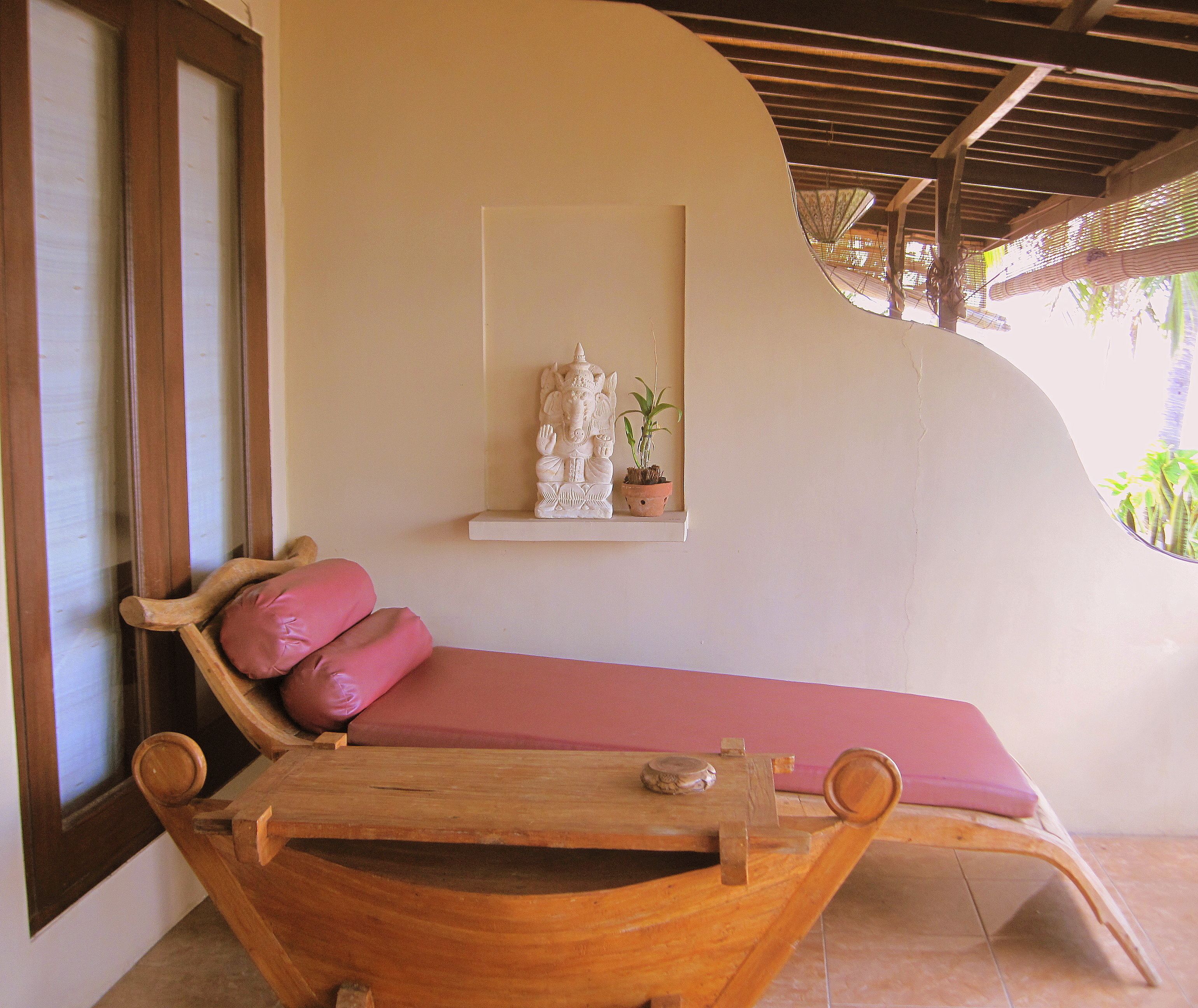 A wooden bench with a pink cushion, a small shelf with a statue and a potted plant, and a window with blinds.
