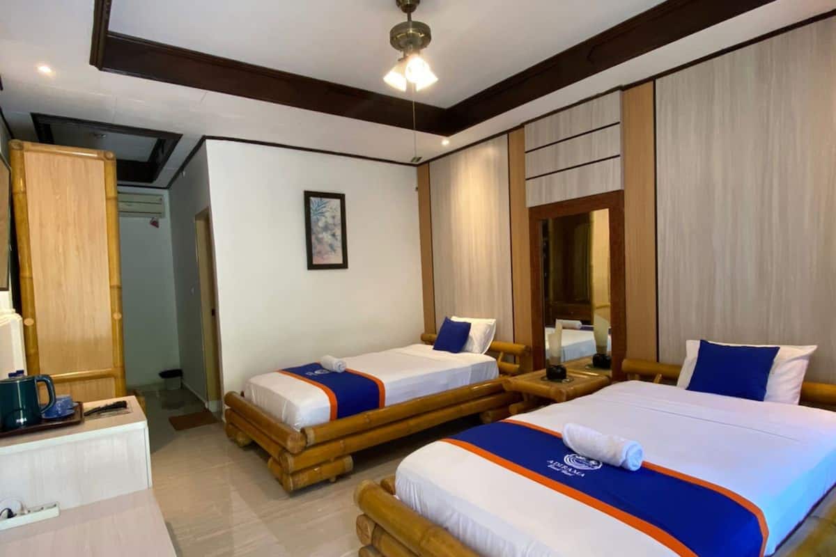 A hotel room with two single beds, wooden headboards, a wooden wardrobe, a bedside table with a lamp, and a small desk with a coffee maker.