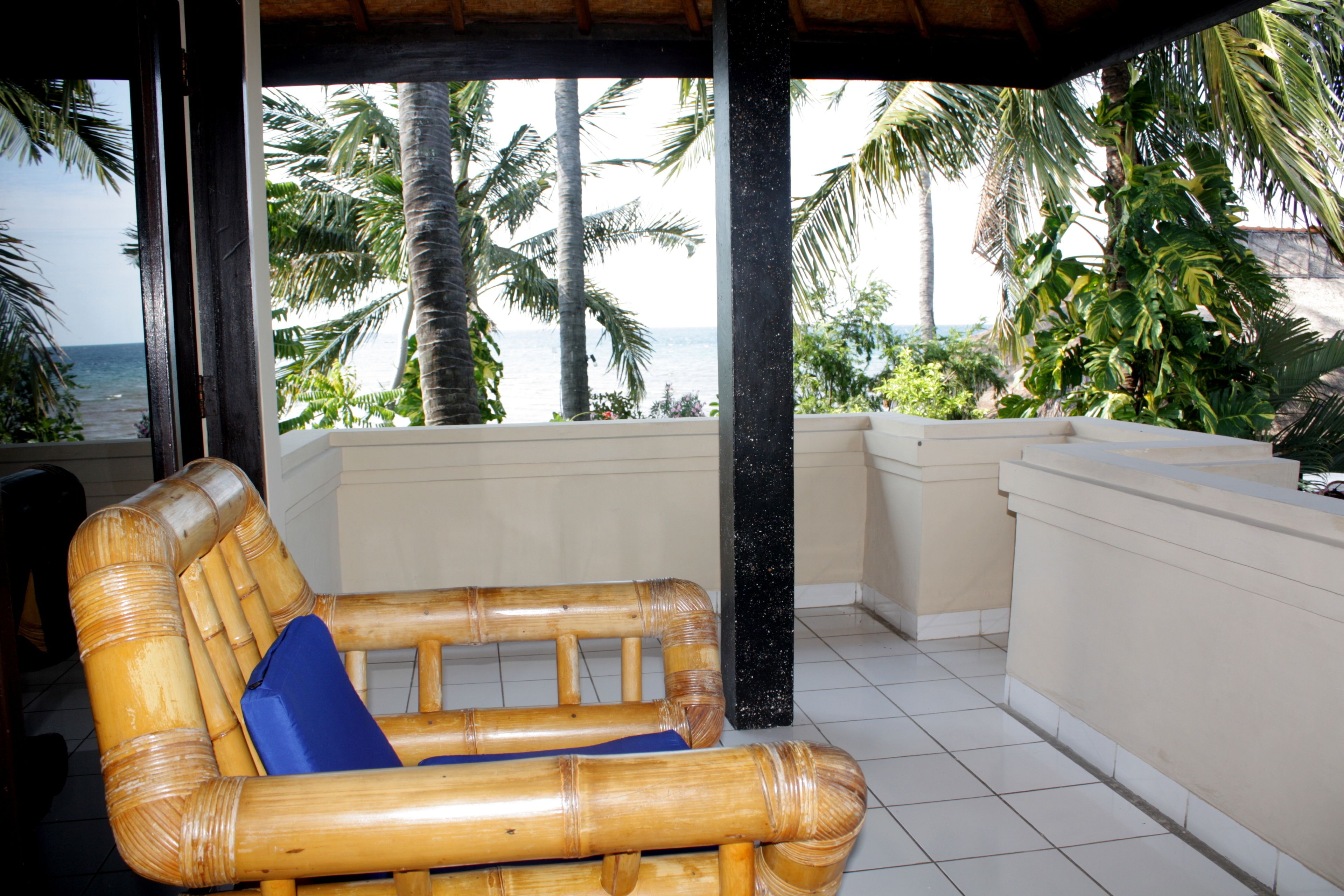 A bamboo sofa with a blue cushion in a covered outdoor area with a view of the ocean and palm trees.
