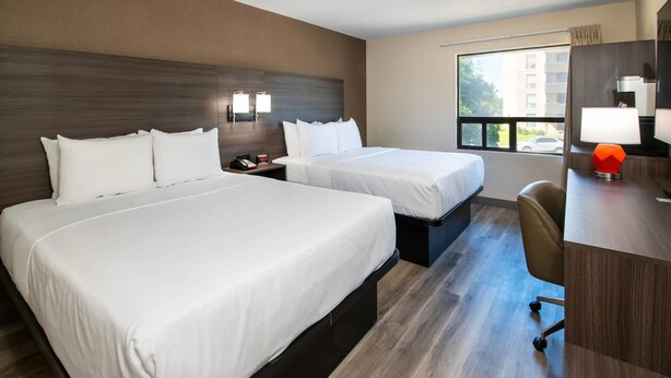 Une chambre d’hôtel avec deux lits, un bureau, une chaise, une table de chevet et une fenêtre donnant sur un immeuble.