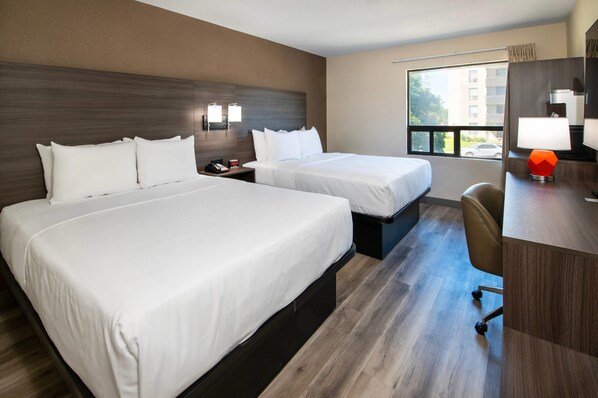 Habitación de hotel con dos camas, un escritorio, una silla, una mesita de noche y una ventana con vista a un edificio.