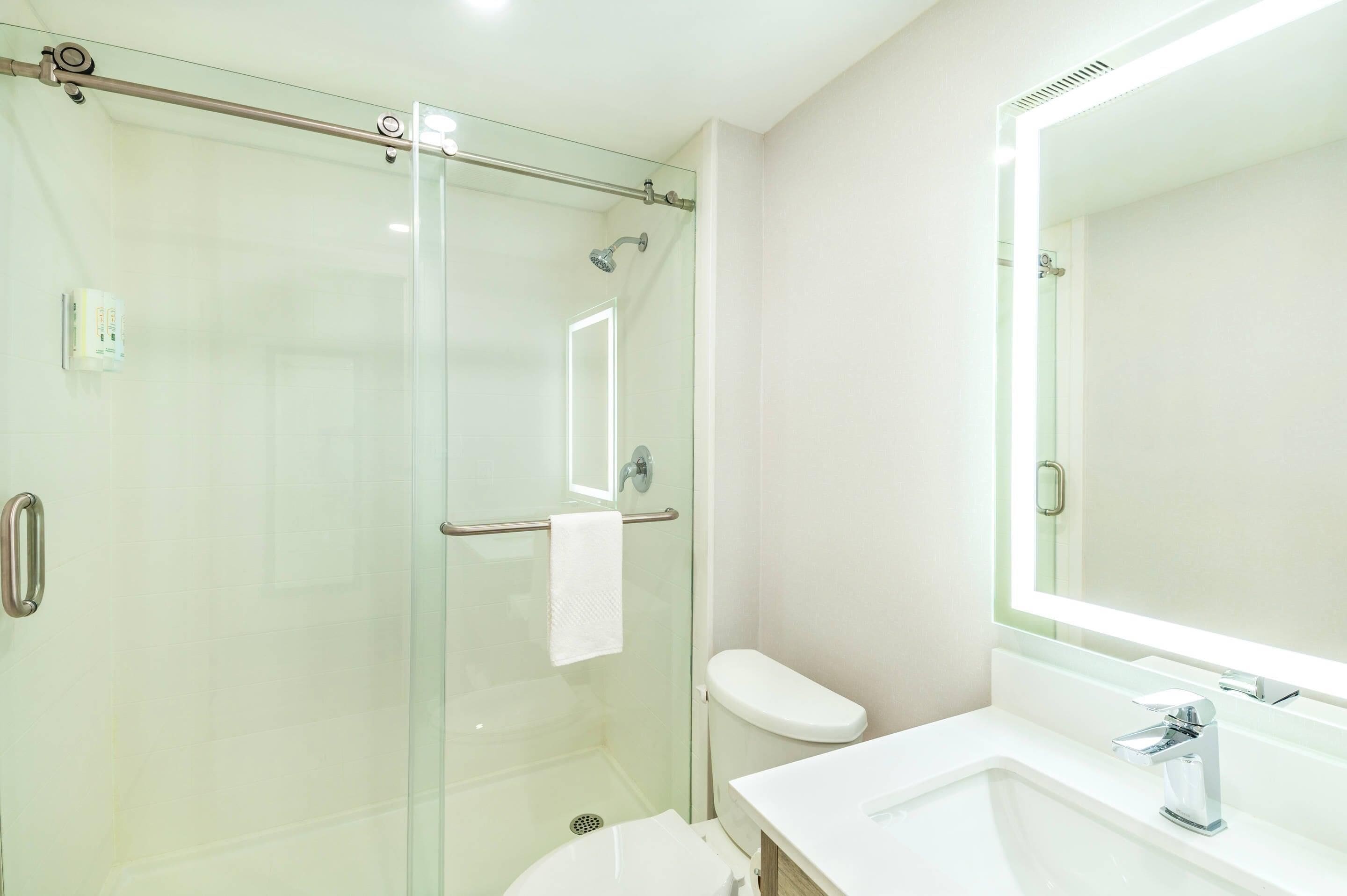 Un baño moderno con cabina de ducha de vidrio, inodoro blanco y un espejo grande sobre el lavamanos.