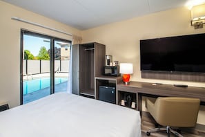 Habitación de hotel con cama, escritorio, televisión, microondas, refrigerador pequeño y vista a una piscina al aire libre.