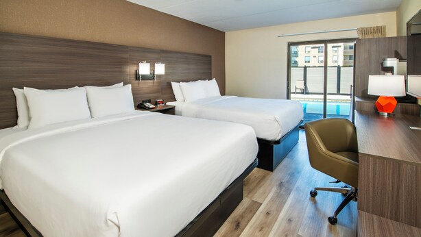 A hotel room with two beds, a desk with a chair, a nightstand, and a window with a view of buildings. - Quality Inn (Chatham-Kent)