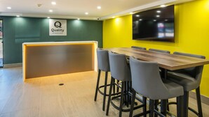 Un espace d’accueil d’hôtel au design moderne, comprenant un comptoir de réception, un coin salon avec une table et des chaises en bois, ainsi qu’un grand téléviseur à écran plat fixé au mur.
