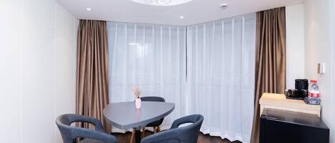 A modern hotel room with a round table, four grey chairs, a small vase with dried flowers, a coffee machine, and a refrigerator.