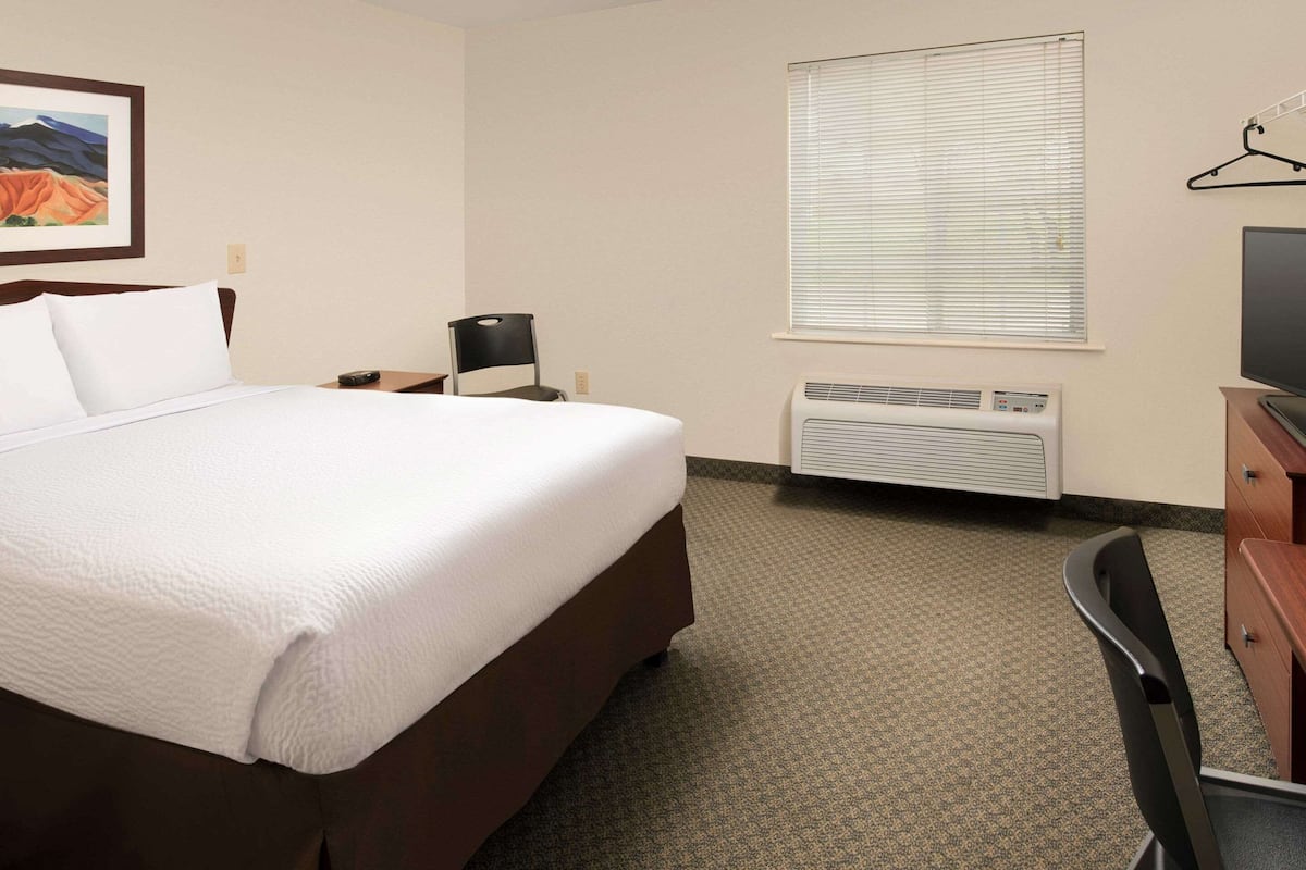 A hotel room with a large bed, a desk, a chair, a TV, an air conditioner, and a window with blinds.