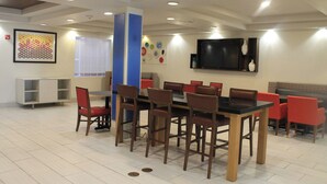 A dining area with a long table and multiple chairs, a television mounted on the wall, and a bookshelf.