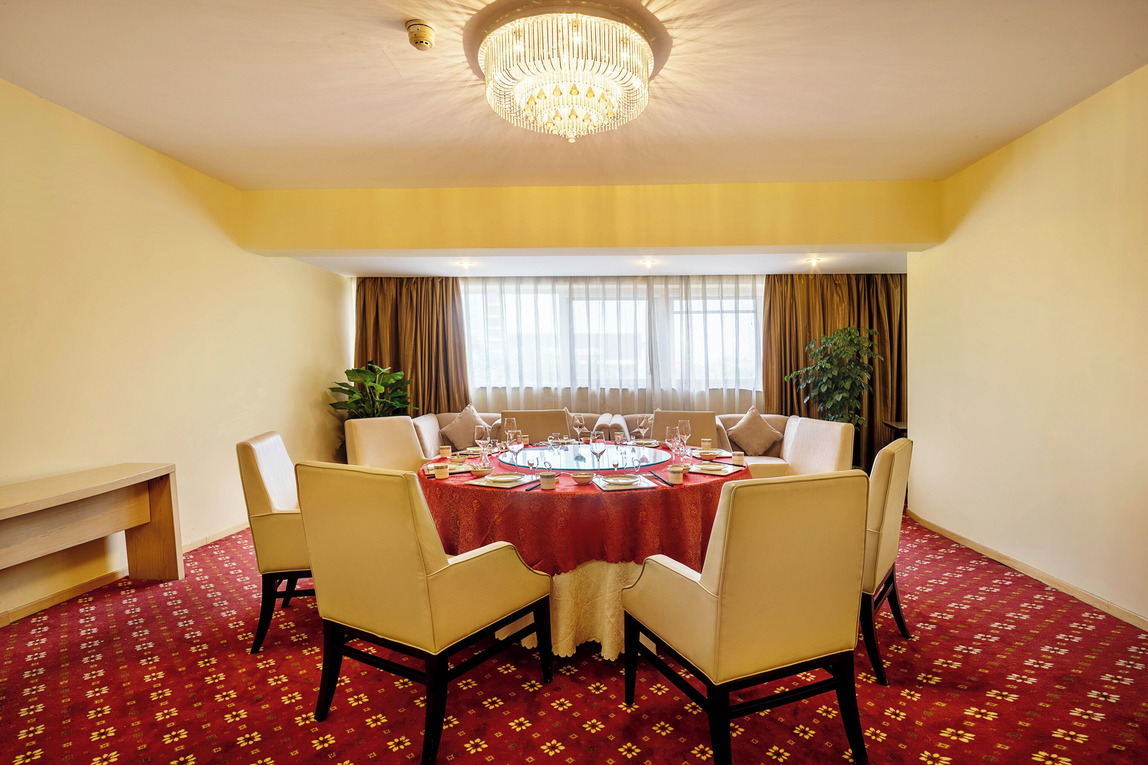 Comedor con una mesa redonda dispuesta para seis comensales, rodeada de seis sillas, una araña y un ventanal con cortinas.