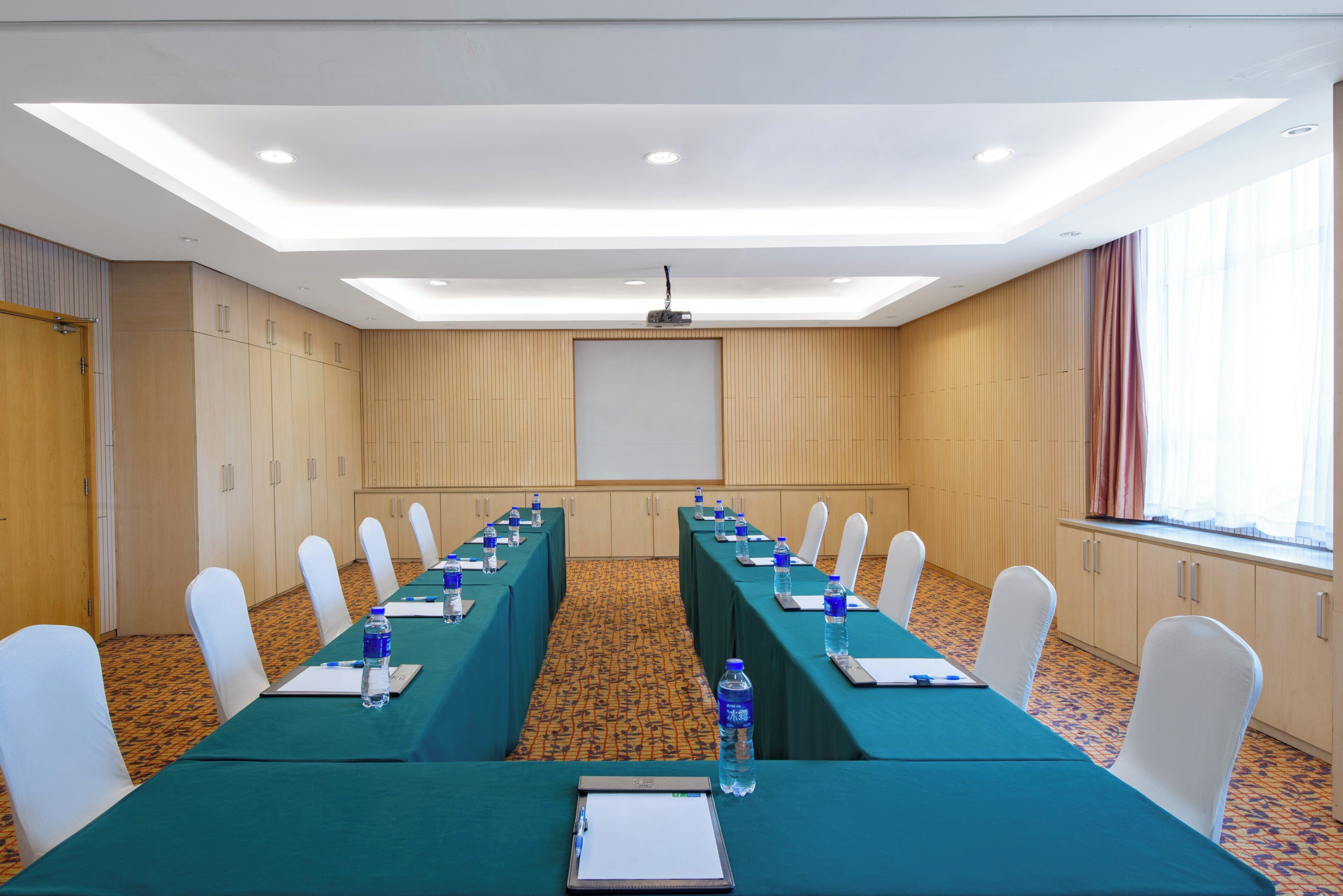 Un salón de conferencias con una larga mesa, sillas, una pantalla de proyector y un amplio ventanal con cortinas.