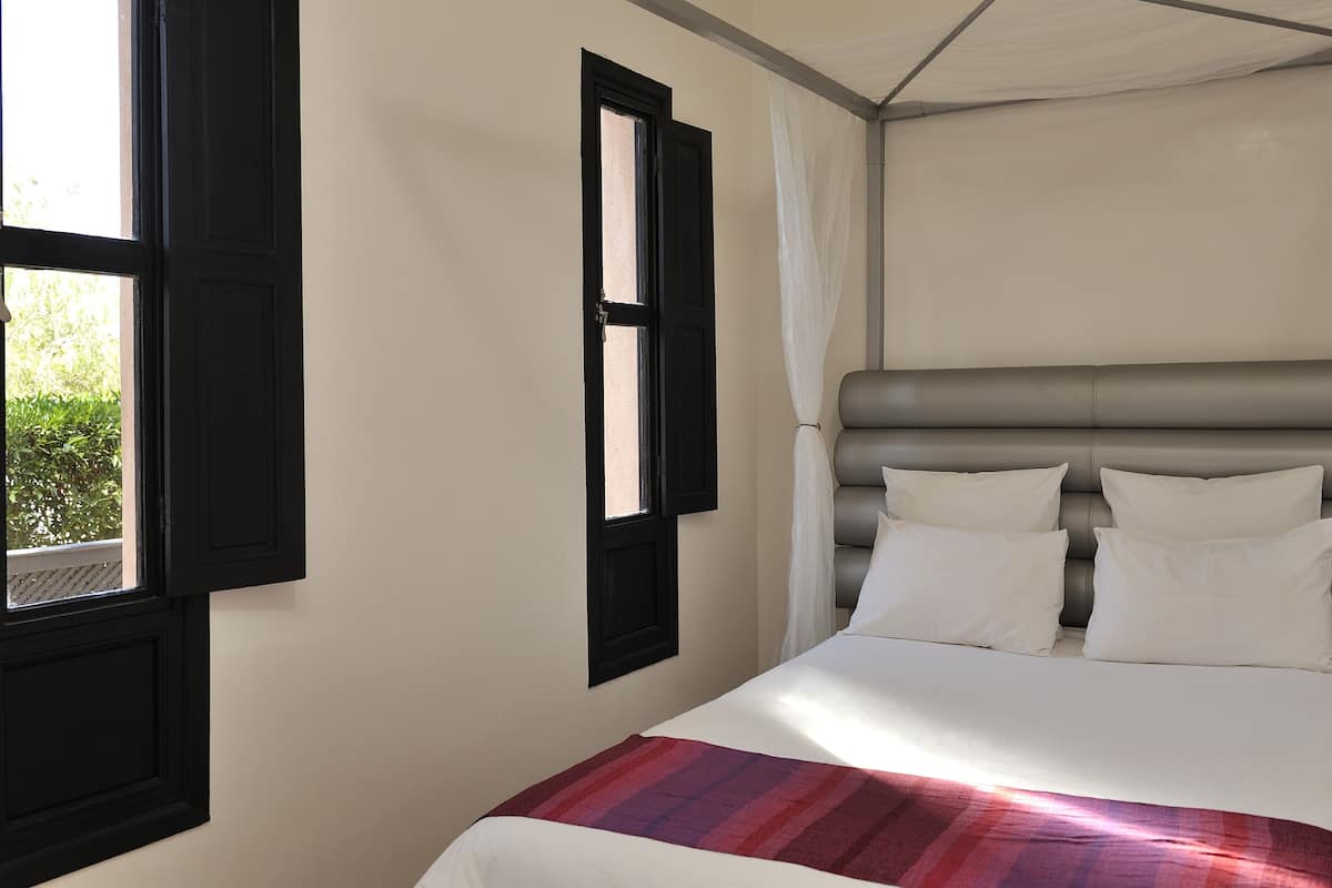 A bedroom with a bed, a window with black shutters, and a white curtain.