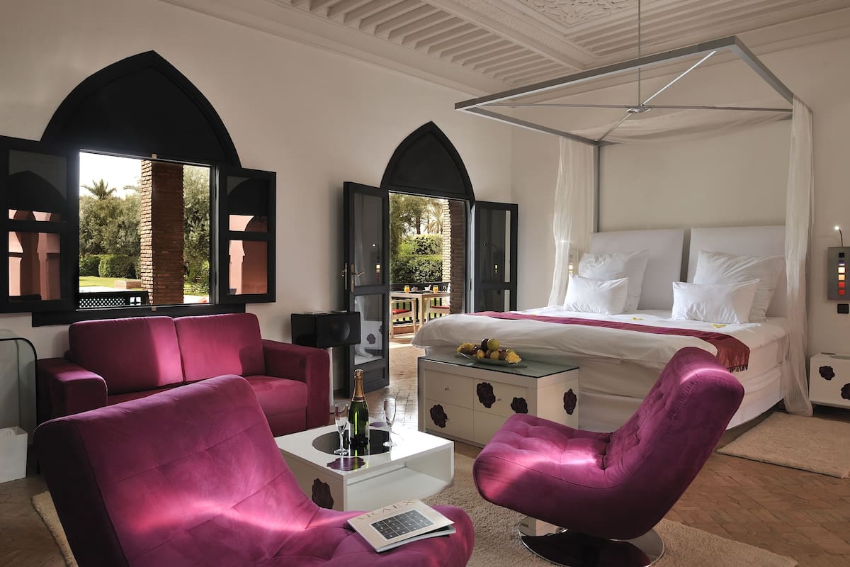 A bedroom with a large bed, a sitting area with a sofa and armchairs, a small table with a bottle and glasses, and a view of the outdoors through arched windows.