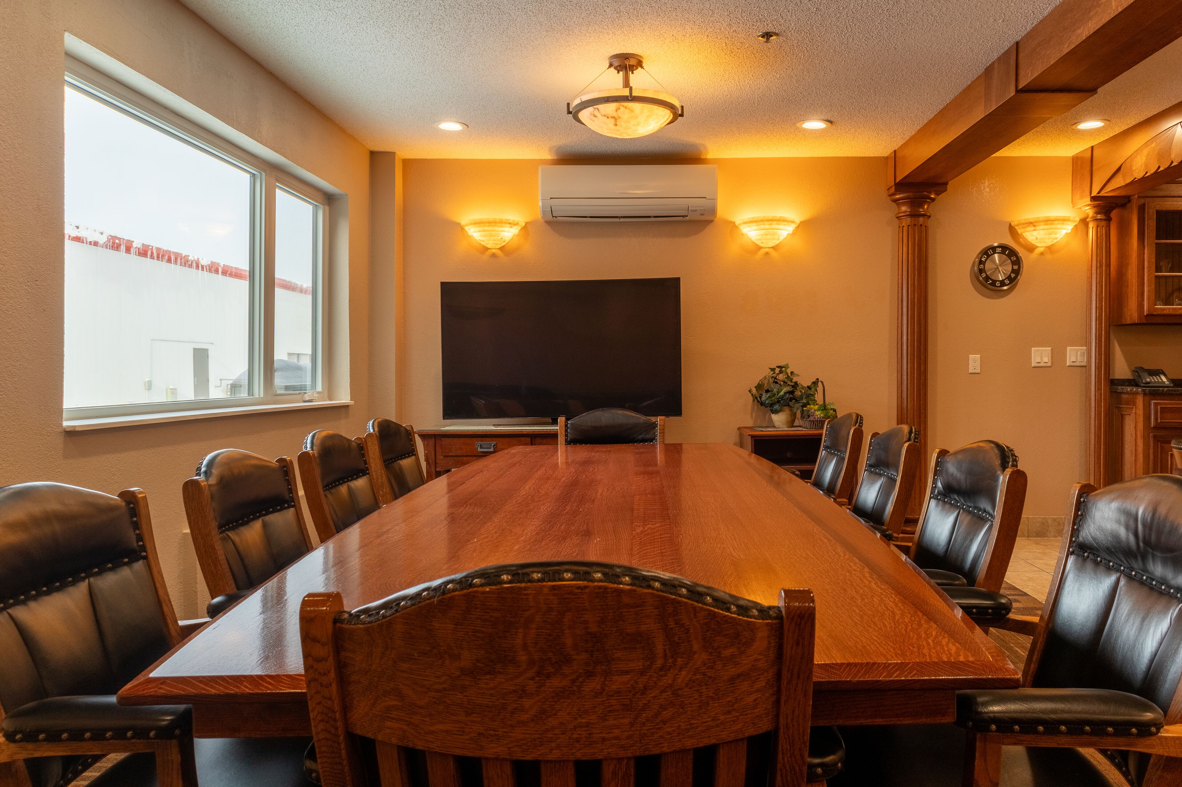 A conference room with a long wooden table, leather chairs, a flat-screen TV, and wall-mounted lighting.