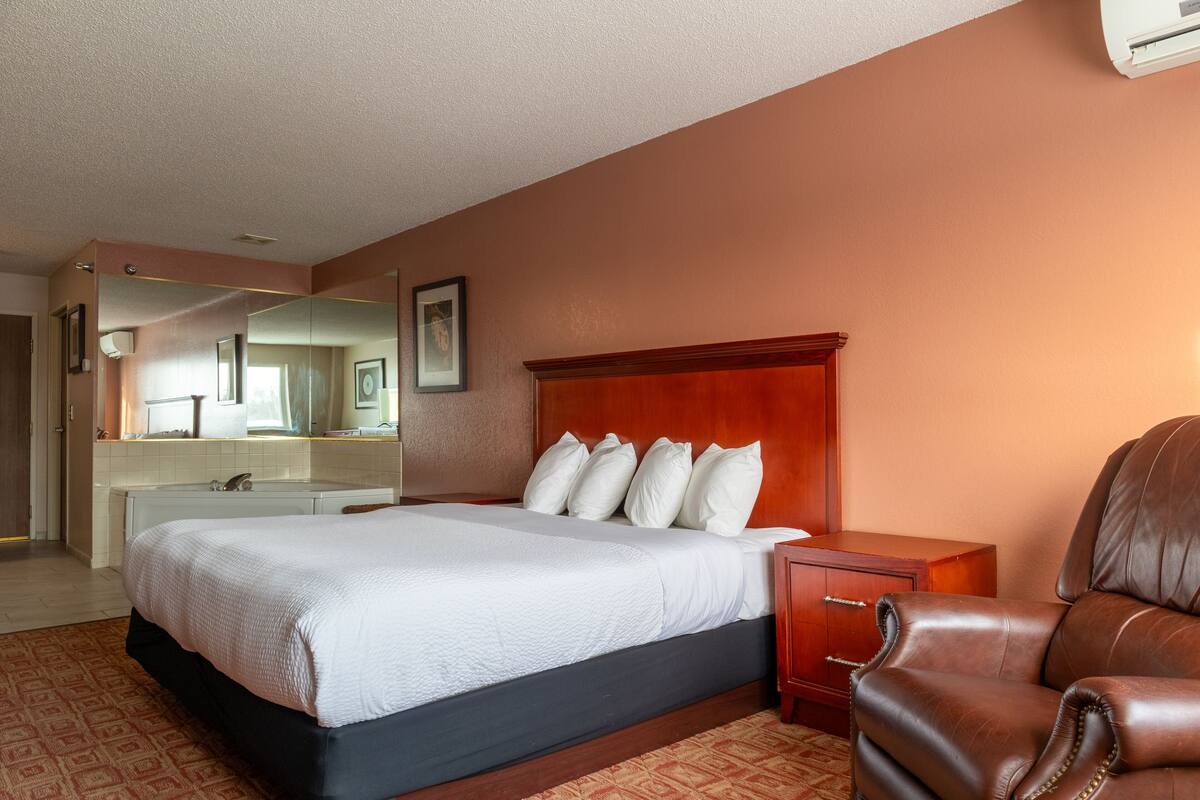 A hotel room with a large bed, a wooden headboard, a brown leather armchair, a wooden nightstand, and a mirror above the bed.