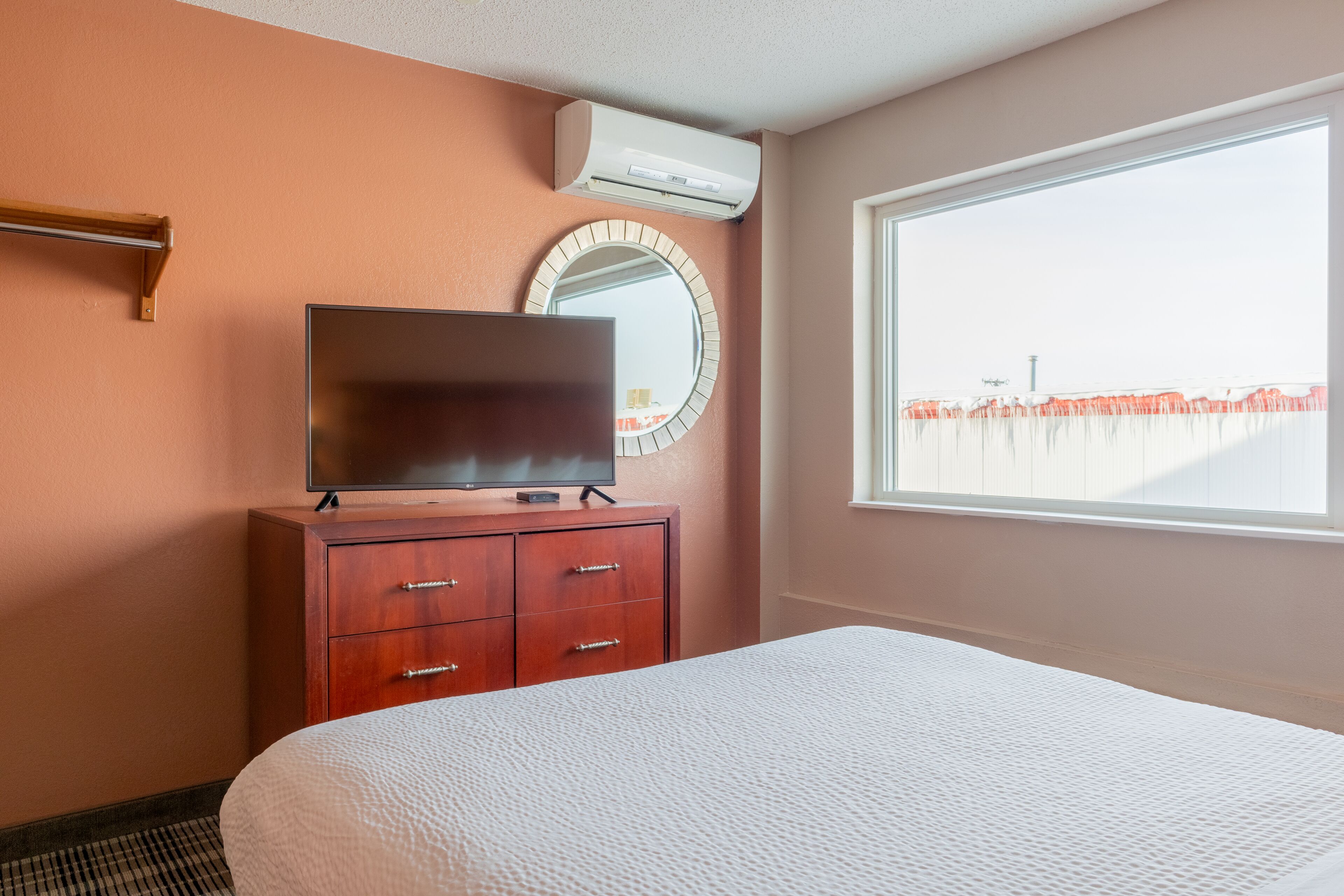 A hotel room with a bed, a TV on a wooden dresser, a round mirror, and a window with a view of a fence.