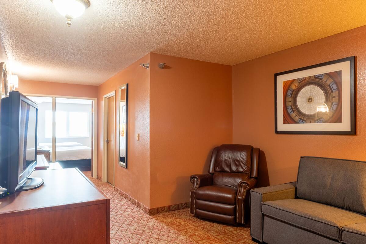 A hotel room with a wooden desk, a leather armchair, a gray sofa, a framed artwork on the wall, and a bed in the background.
