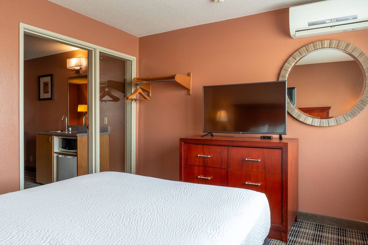 A hotel room with a bed, a wooden dresser, a flat-screen TV, a round mirror, and a kitchenette in the background.