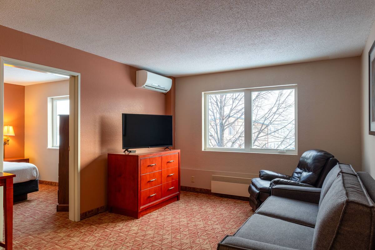 A hotel room with a TV on a wooden dresser, a leather sofa, a window with a view of trees, and a door leading to another room.