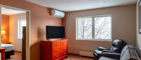 A hotel room with a TV on a wooden dresser, a leather sofa, a window with a view of trees, and a door leading to another room.