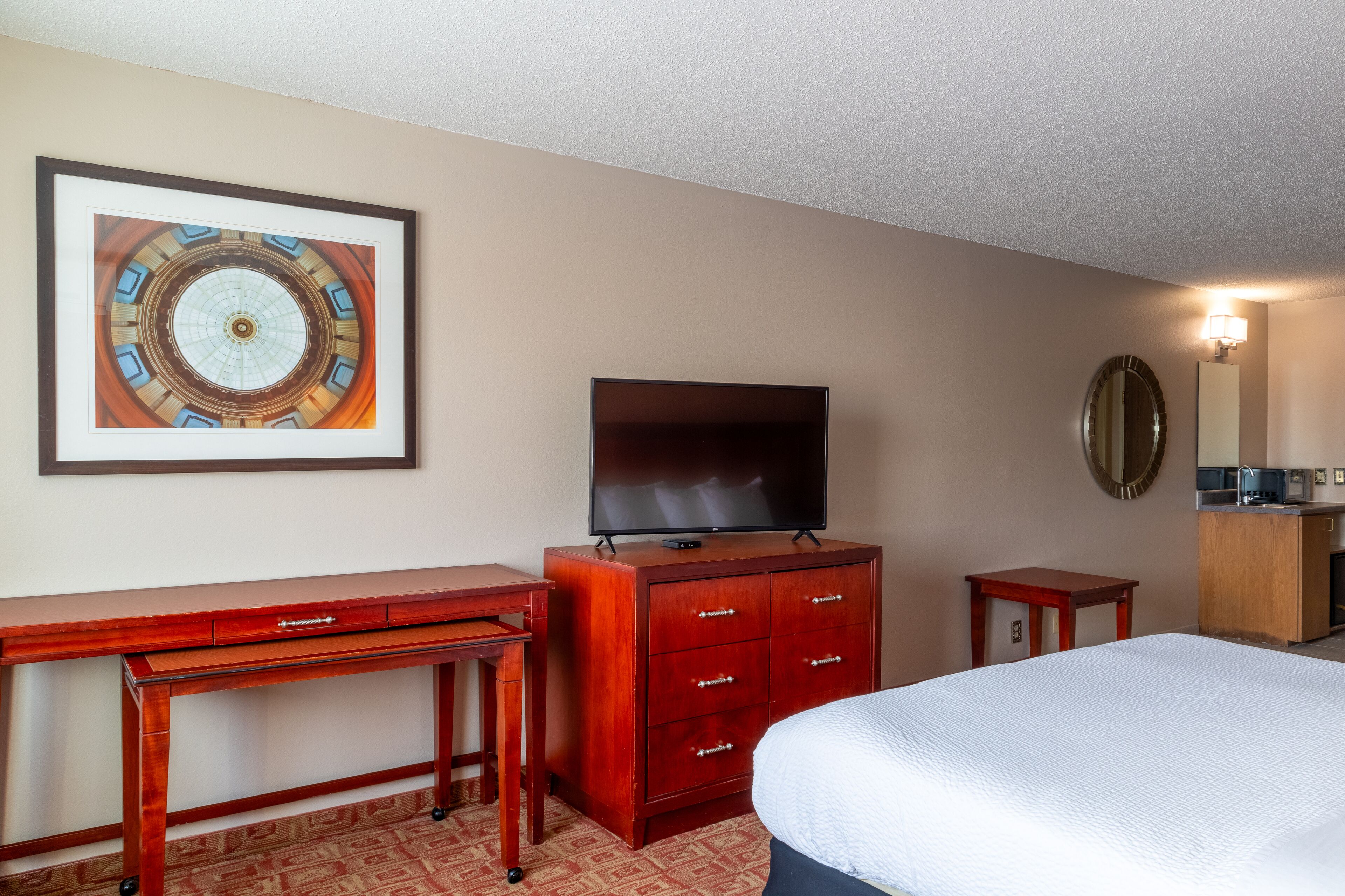A hotel room with a bed, a TV, a wooden dresser, a desk, and a framed picture on the wall.