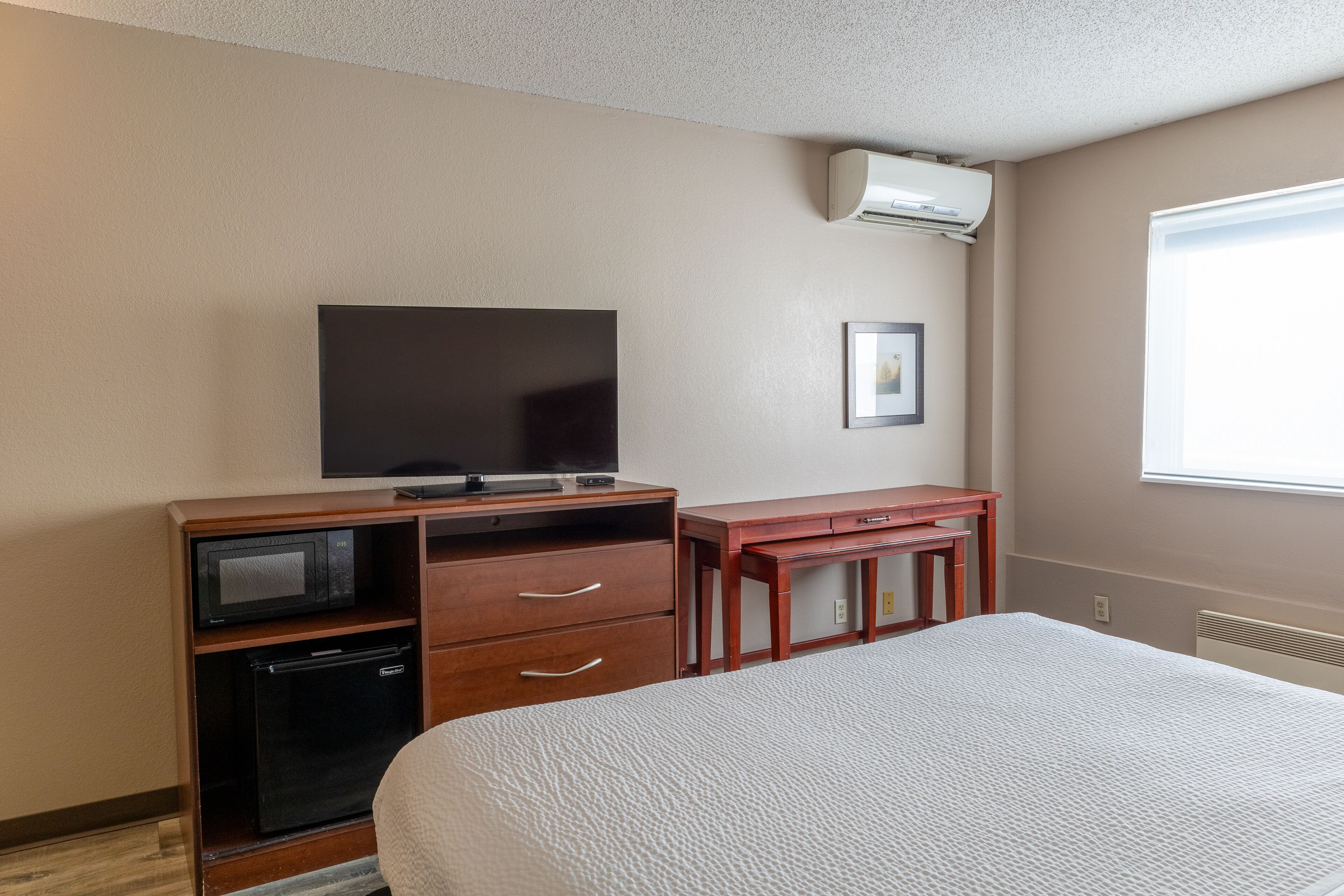 Une chambre d’hôtel équipée d’un lit, d’une télévision, d’un petit réfrigérateur, d’un four à micro-ondes, d’un bureau en bois et d’une fenêtre avec des stores.
