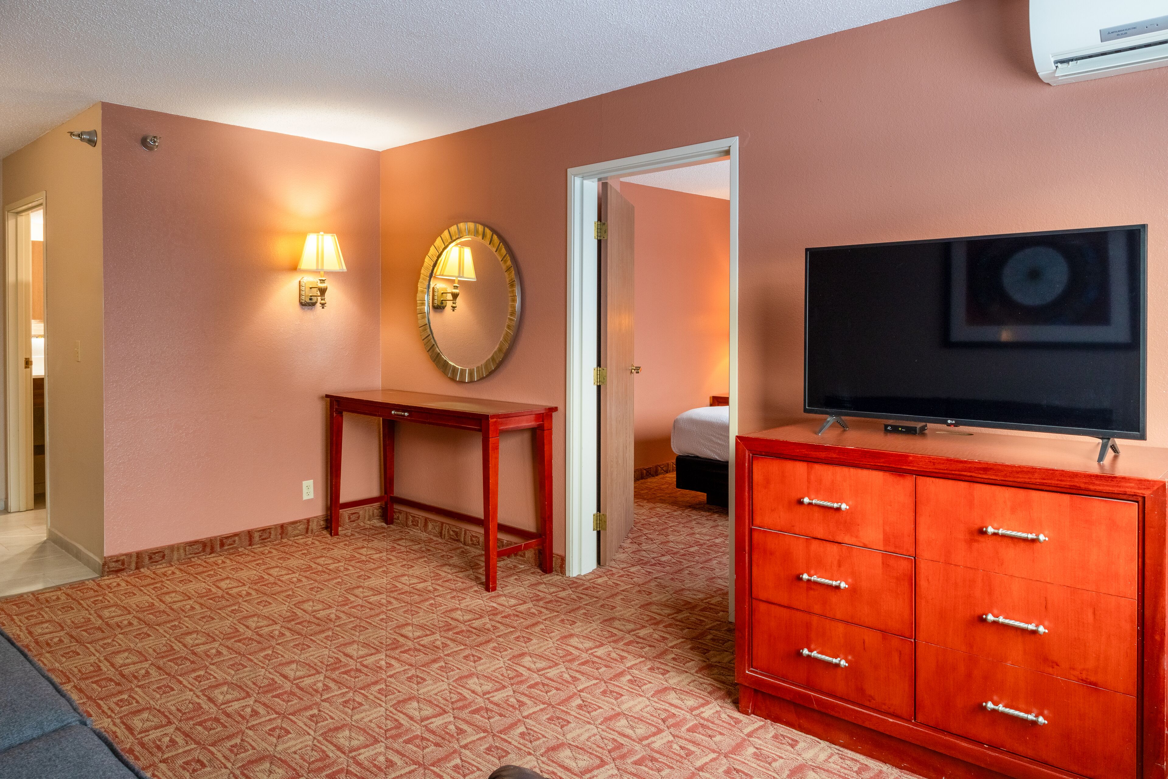Una habitación de hotel con alfombra estampada, cómoda de madera con cajones, televisor, espejo redondo con luz y una puerta que da a otra habitación.
