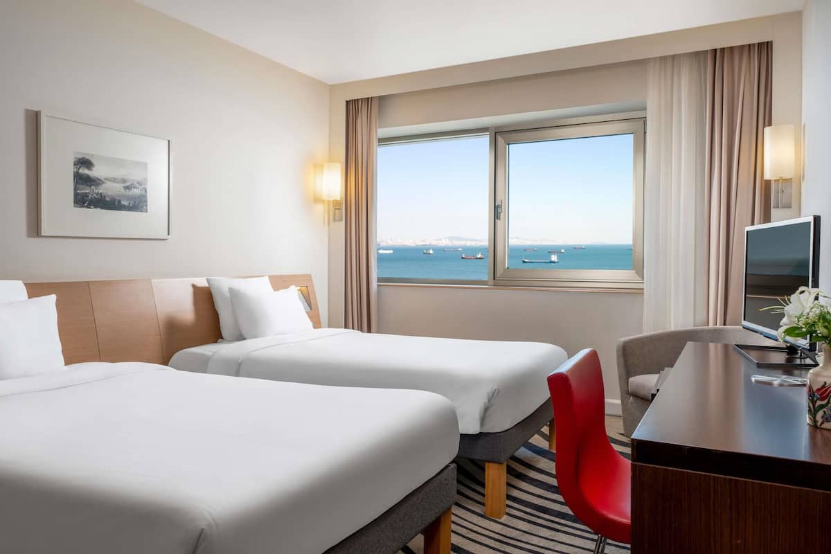 A hotel room with two beds, a desk with a TV, a chair, a window with a view of the sea, and a framed picture on the wall.