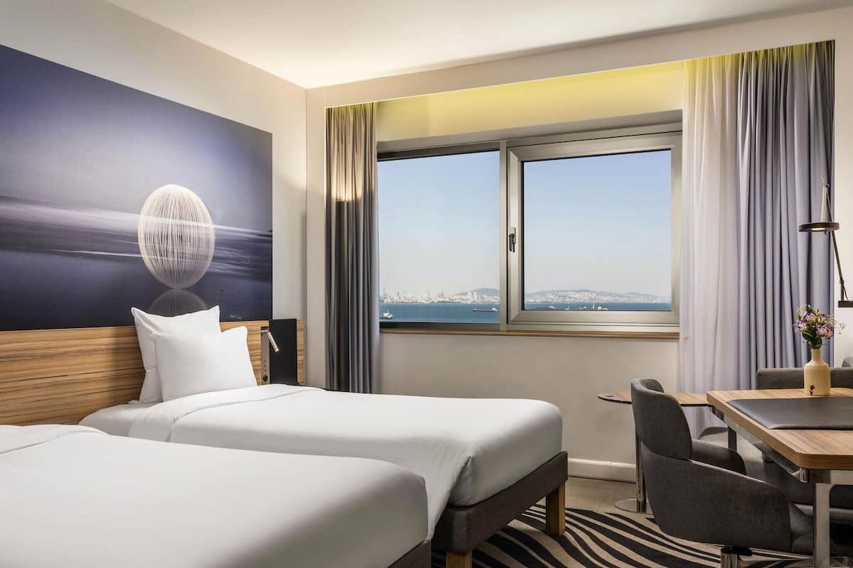 Hotelkamer met twee bedden, een bureau, een stoel, stadszicht en een groot raam.