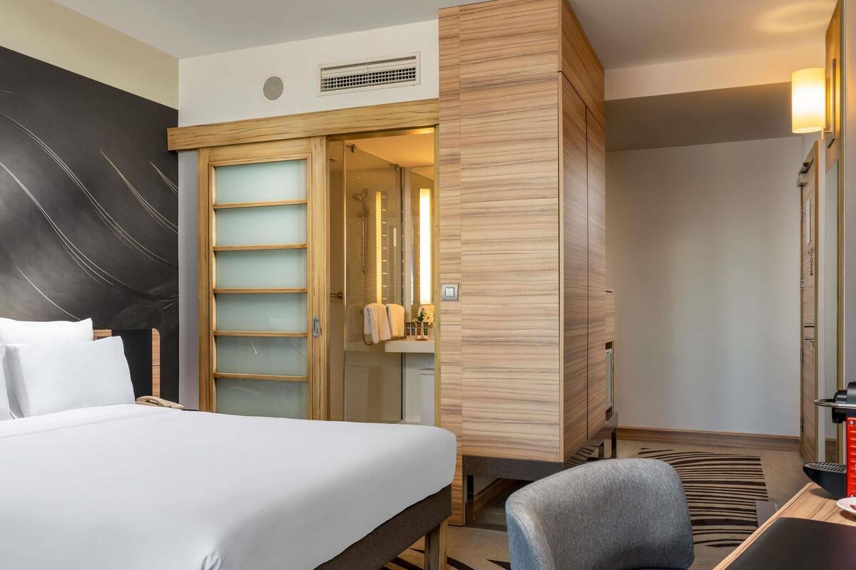 A hotel room with a bed, a desk, a chair, a wardrobe, and a bathroom visible through an open door.