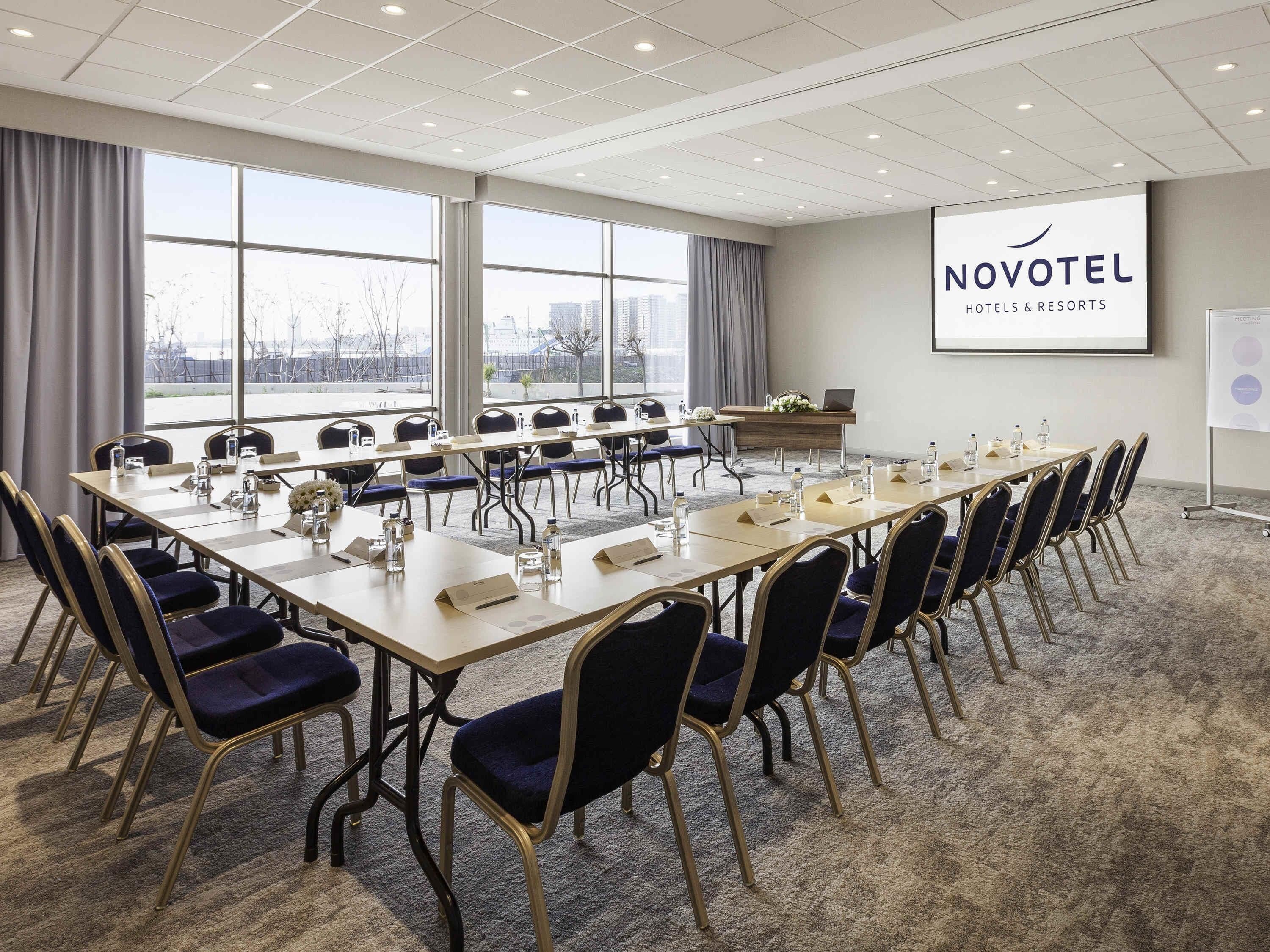 A conference room with a long table, chairs, a projector screen, and a view through large windows.