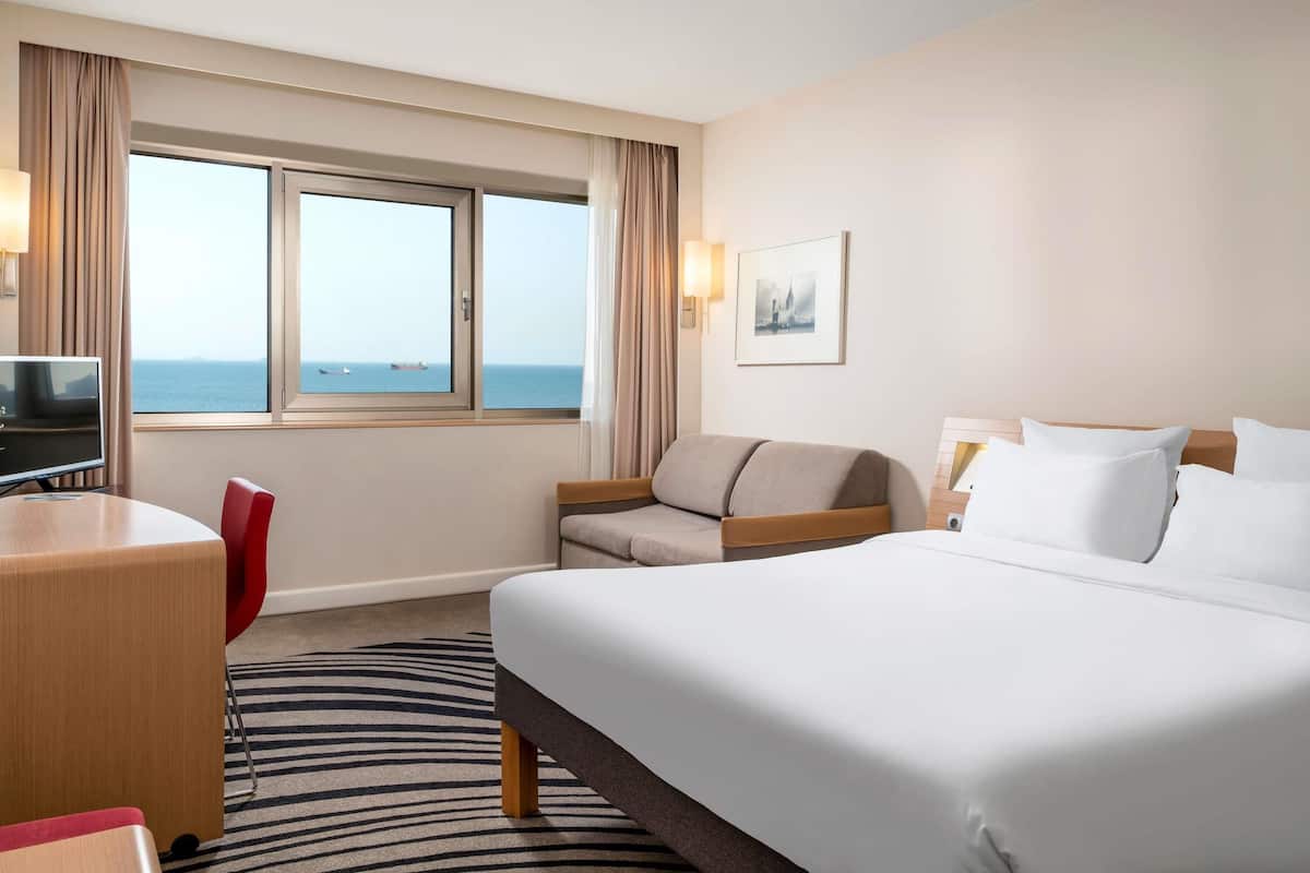 A hotel room with a bed, a desk, a chair, a television, and a window with a view of the sea.