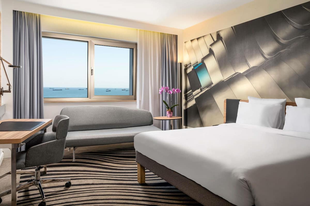 A hotel room with a bed, a desk, a chair, a view of the sea, and a modern headboard.
