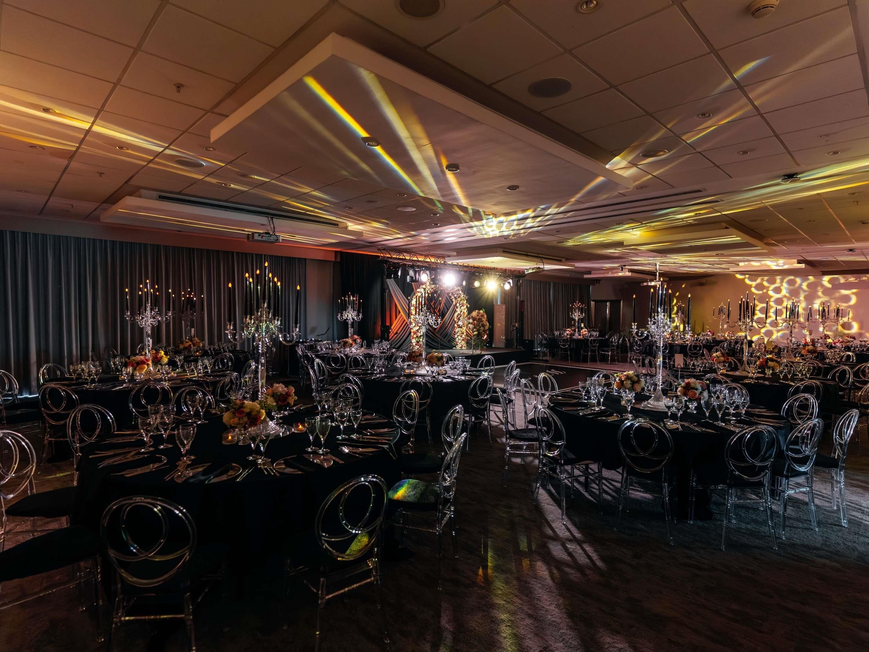 Un salón de banquetes con varias mesas redondas, cada una con cristalería y cubiertos, y un escenario decorado con arreglos florales.