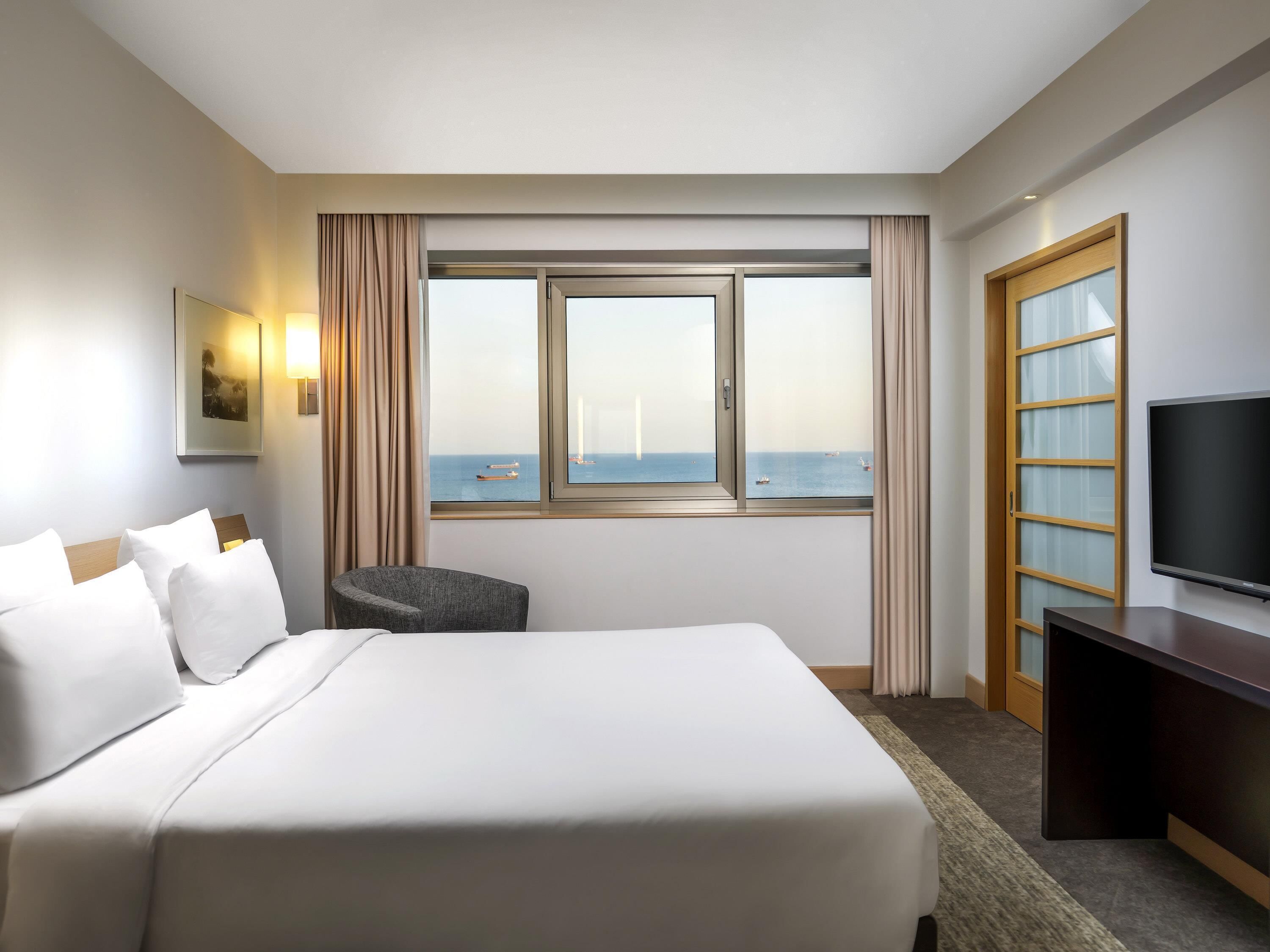 A hotel room with a large bed, a chair, a TV, and a window with a view of the sea.