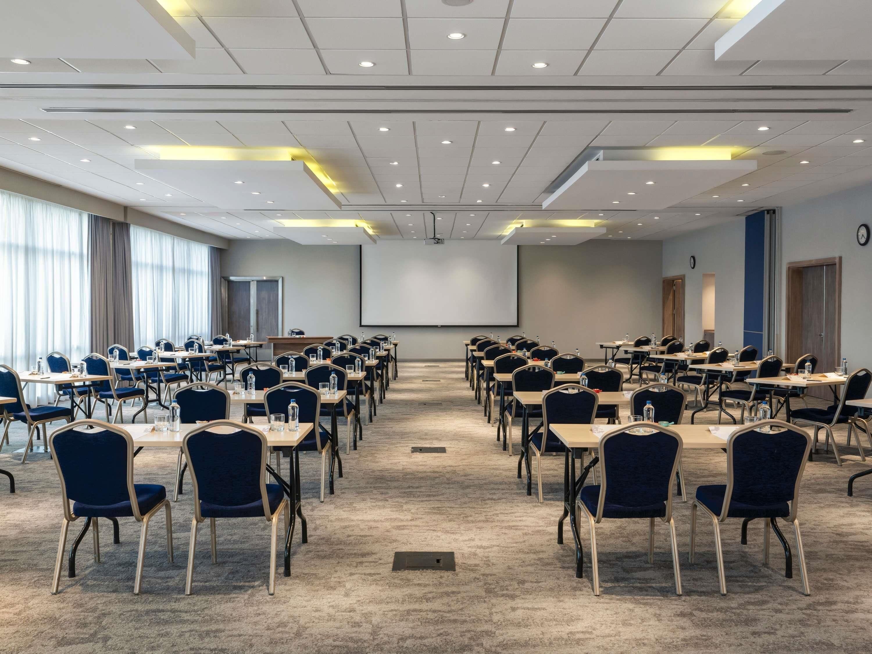A conference room with rows of tables and chairs, a projector screen, and a large window with curtains.