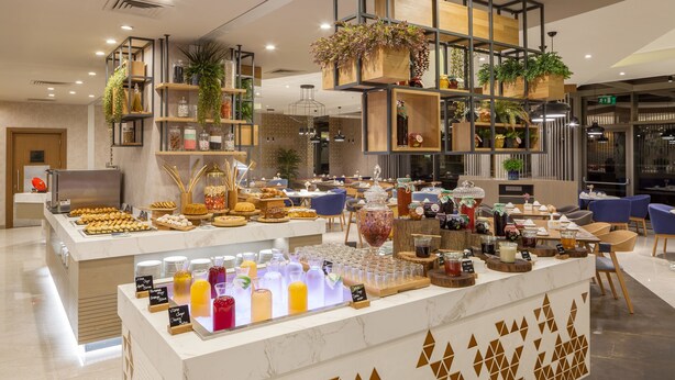 A modern hotel lobby with a central buffet area featuring a variety of food and drink options, surrounded by seating and decorative plants. - Novotel Istanbul Zeytinburnu (Istanbul)