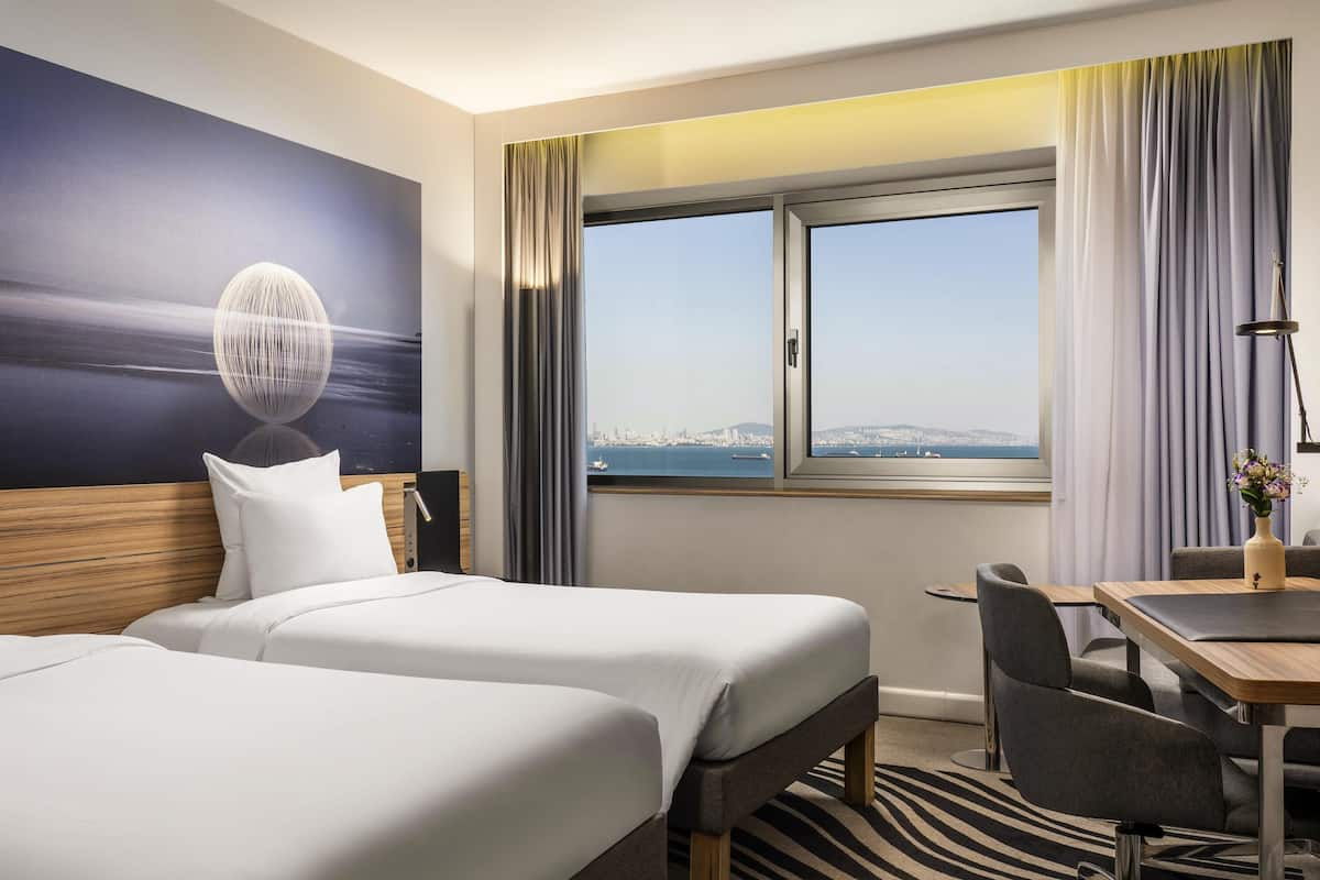 A hotel room with two beds, a desk, a chair, a view of the cityscape, and a large window with curtains.