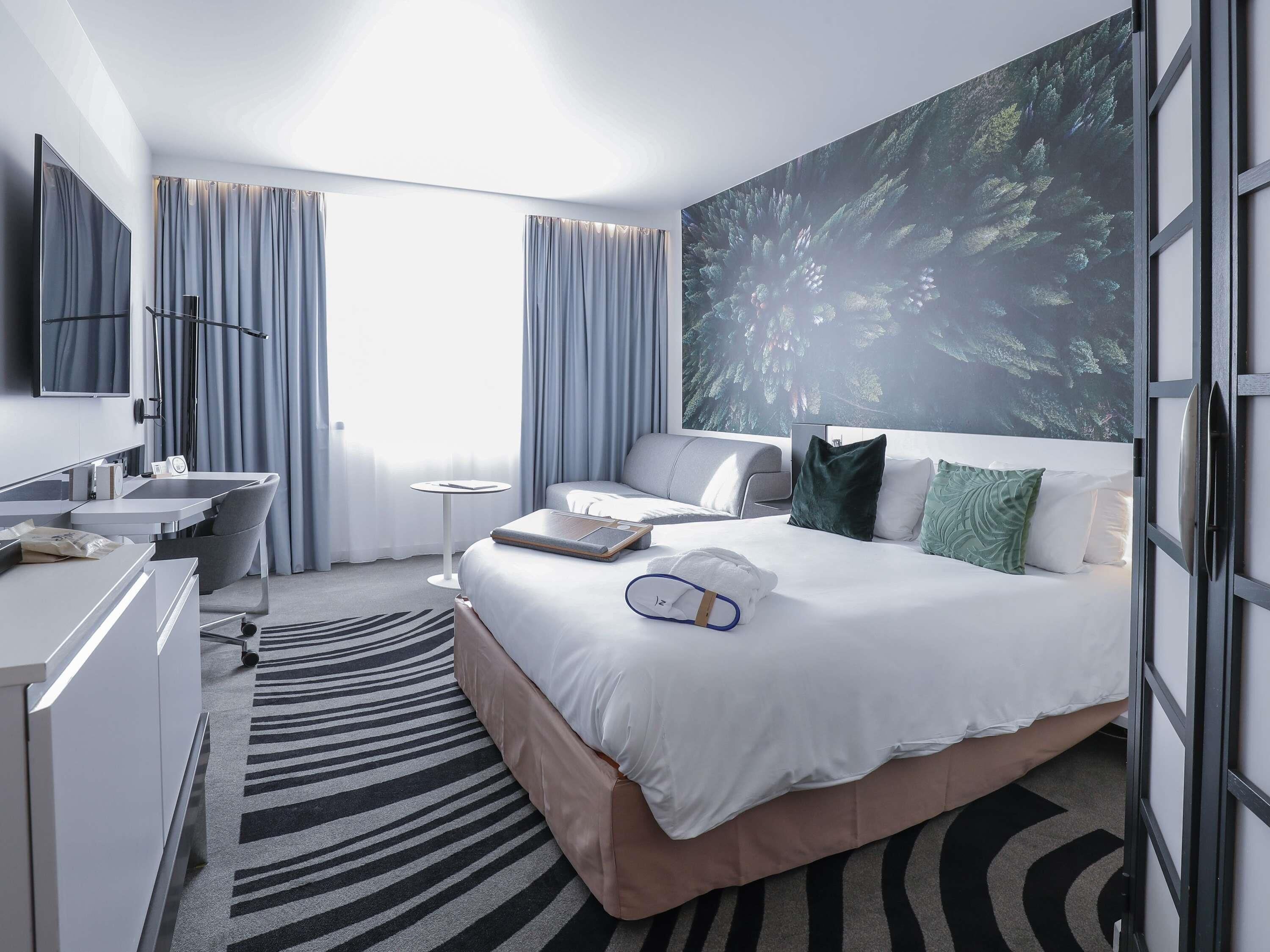 A hotel room with a bed, a desk, a chair, a small table, and a large window with curtains.