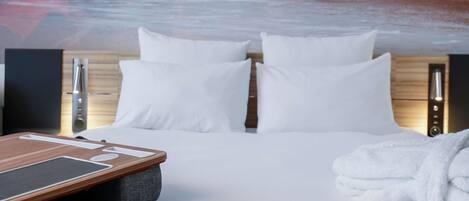 Habitación de hotel con cama, almohadas blancas, cabecero de madera, camilla de masaje y una toalla.