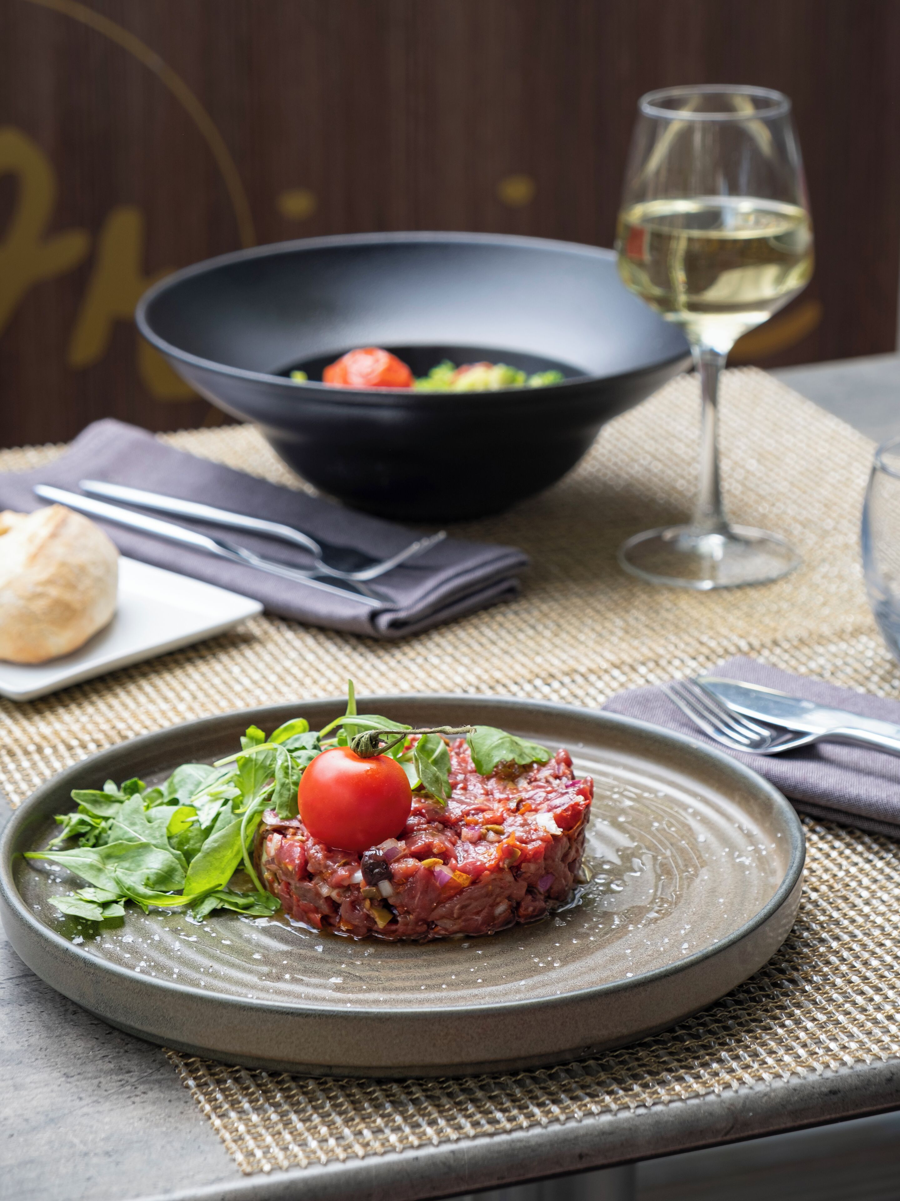 Une assiette de steak tartare garnie de tomates cerises et de jeunes pousses, un saladier rempli de salade et un verre de vin blanc, le tout posé sur une table.