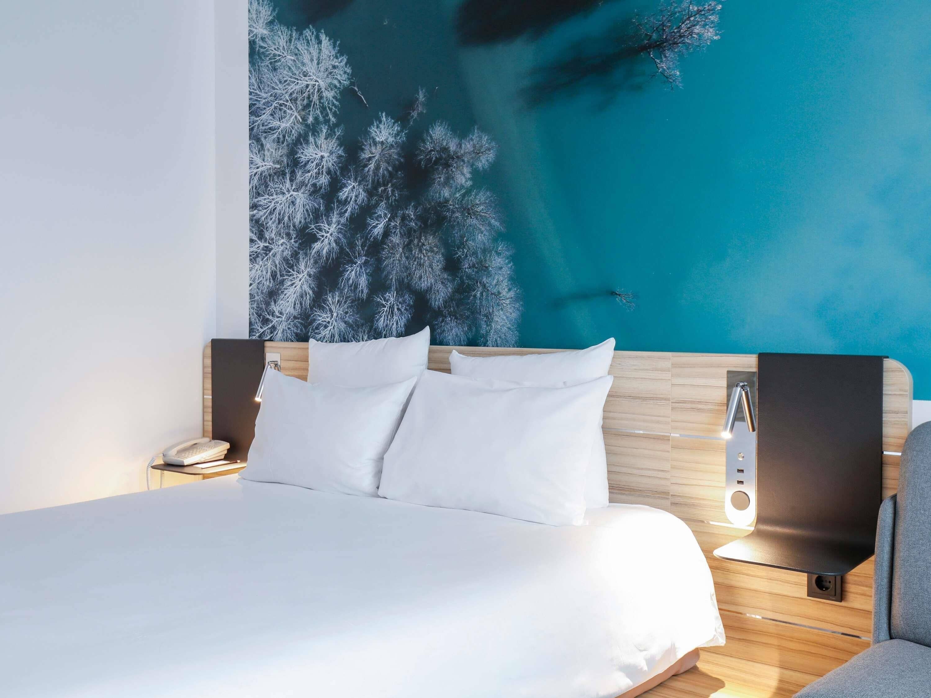 Una habitación de hotel moderna con una cama grande, una mesita de noche, una lámpara de pared y un amplio ventanal con vista a árboles.