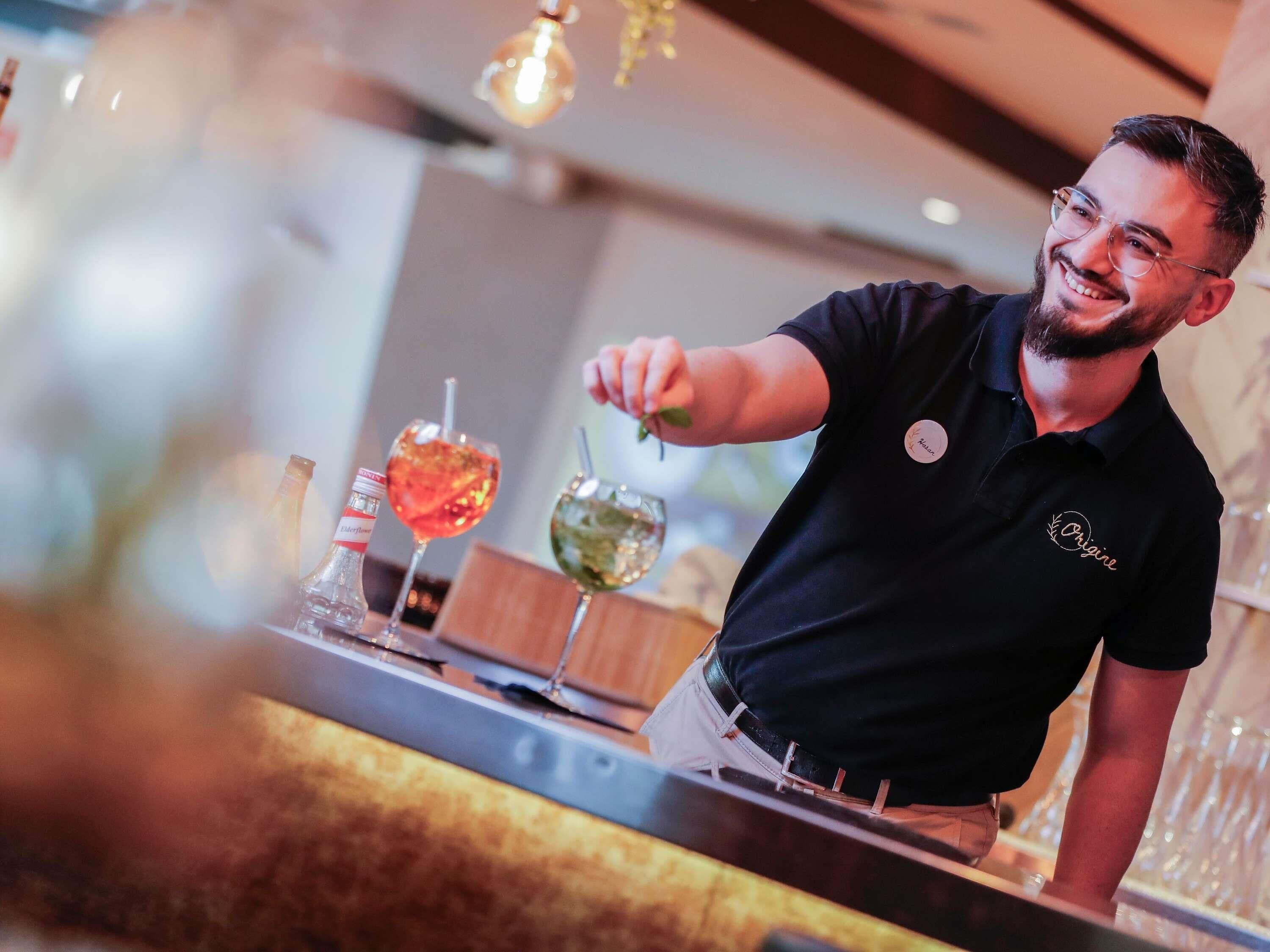A bartender in a black shirt with a badge, preparing drinks behind a bar with two glasses of cocktails and a bottle of liquor.