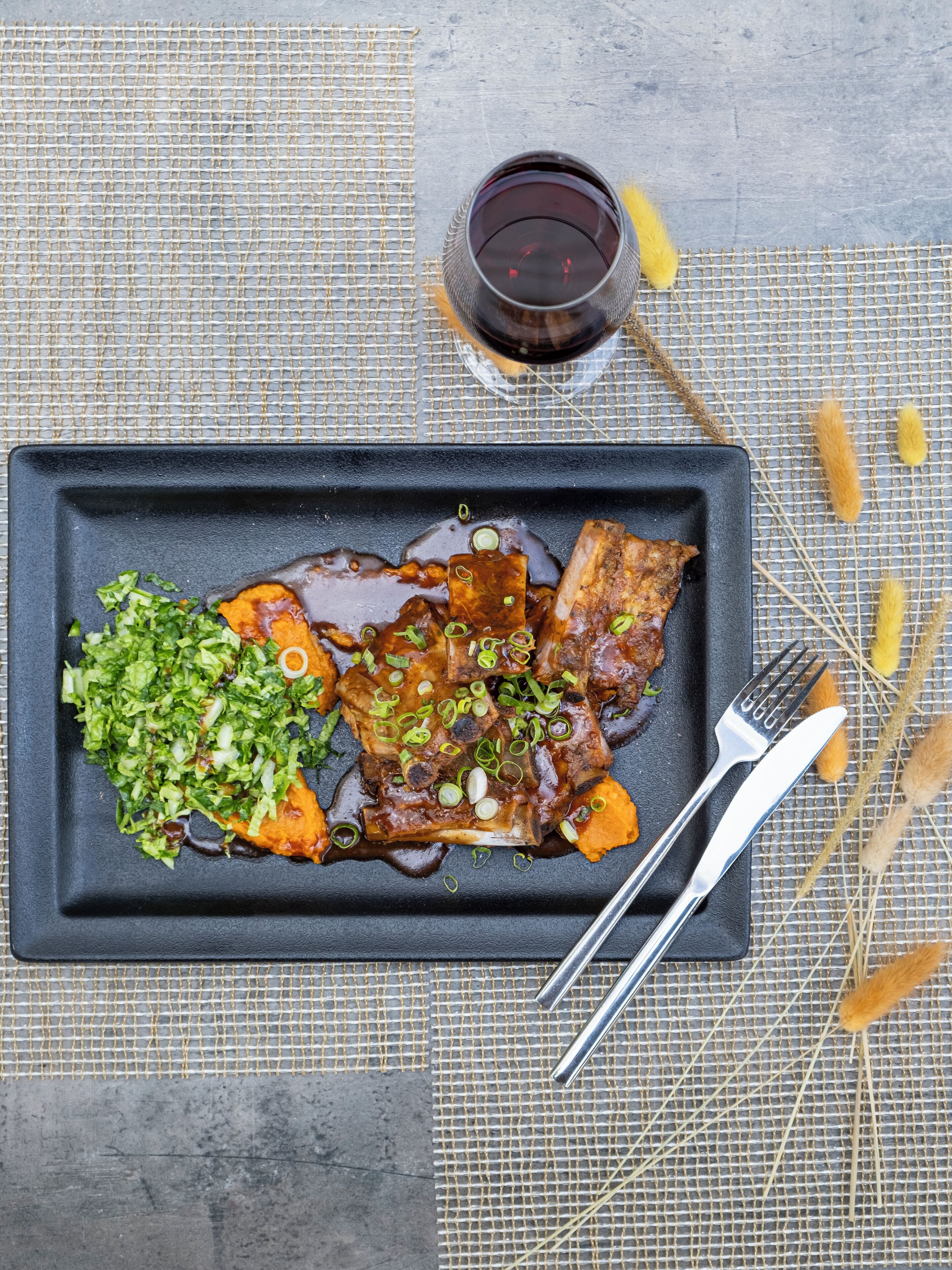 A rectangular plate with a dish of glazed meat and vegetables, a glass of red wine, and a fork and knife on a textured tablecloth.