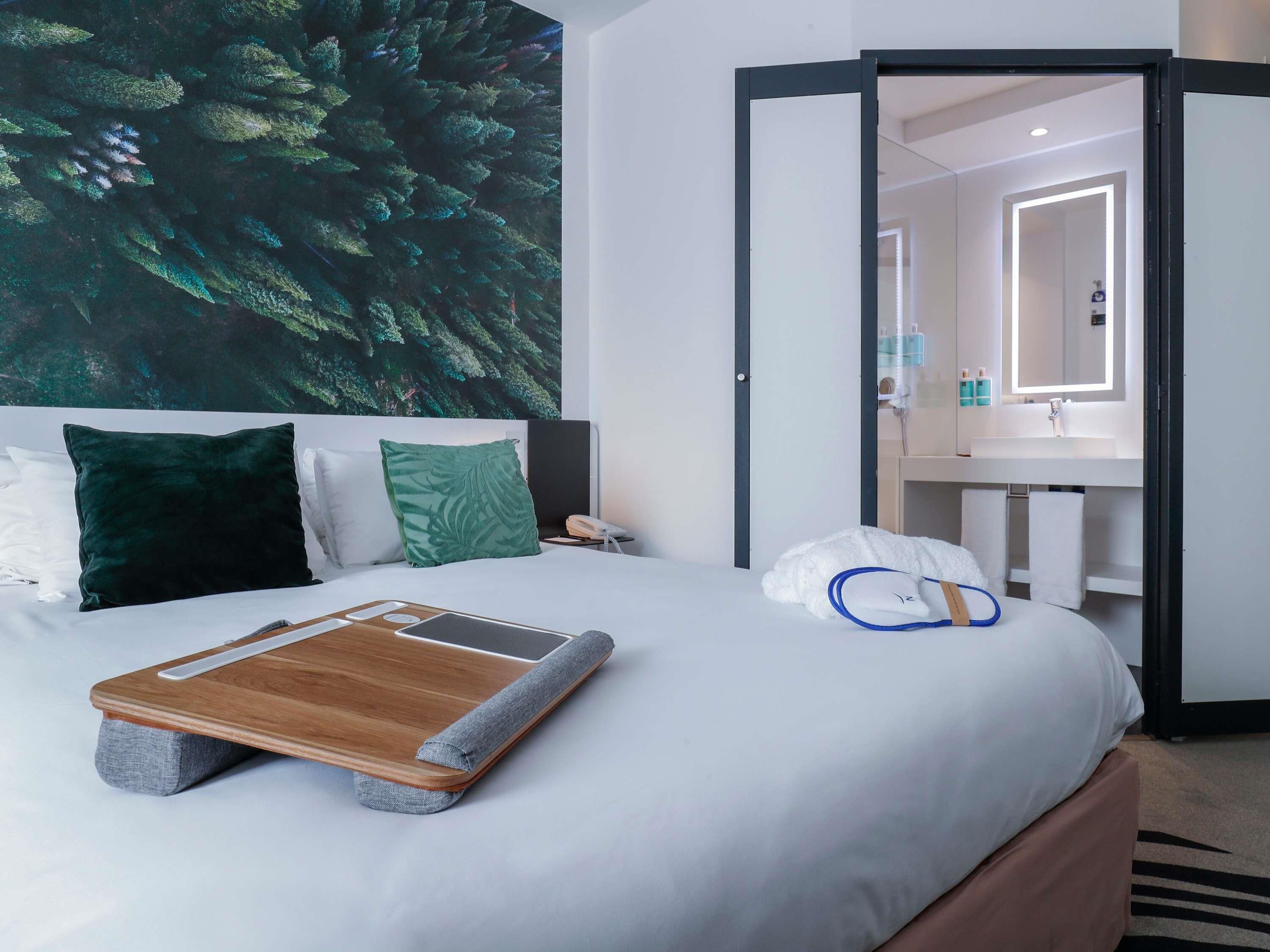 Una habitación de hotel con una cama, una mesita de noche, un teléfono en la pared, un baño con espejo y una toalla en el suelo.