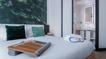 A hotel room with a bed, a nightstand, a wall-mounted phone, a bathroom with a mirror, and a towel on the floor.