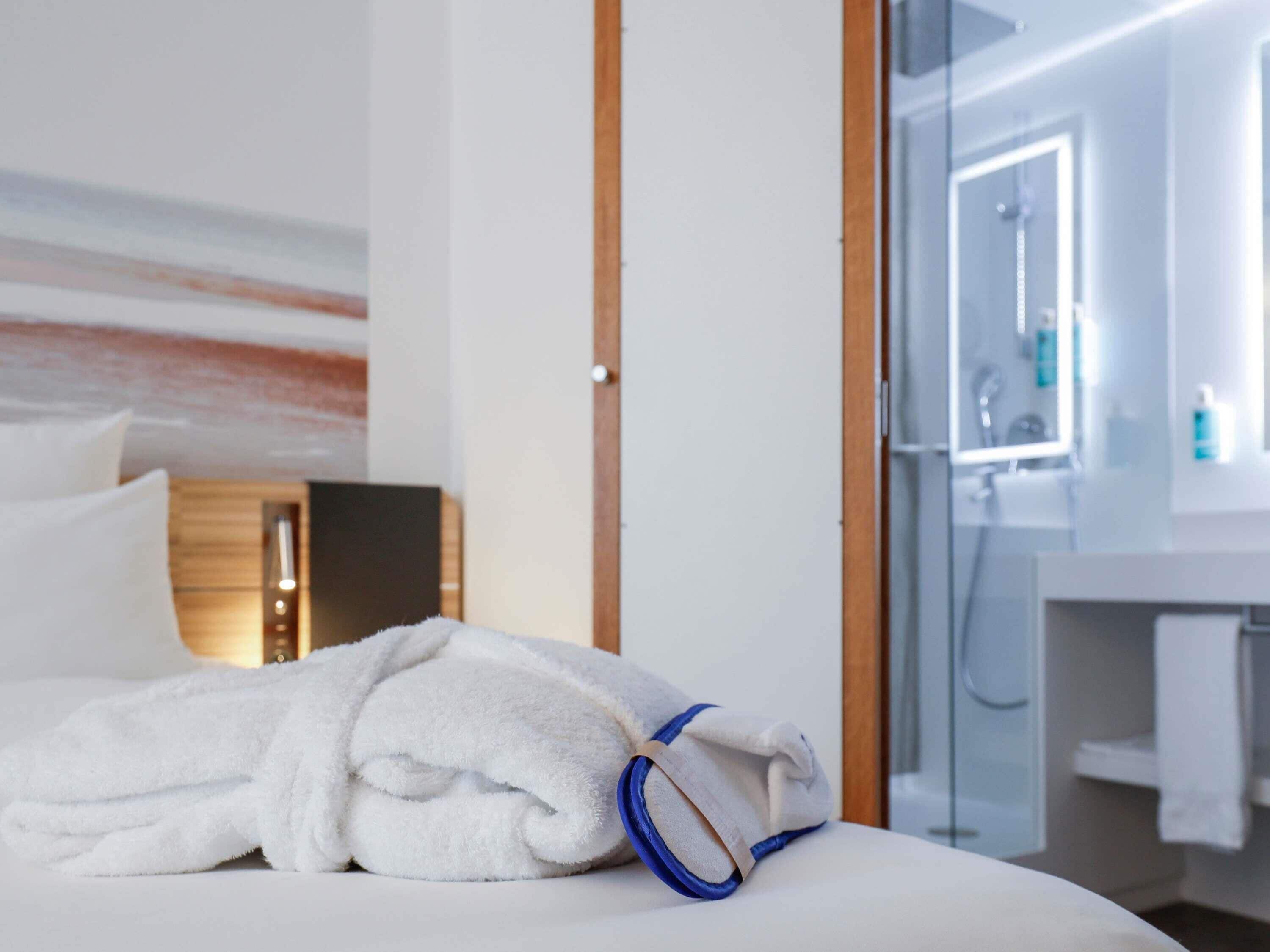 A neatly made bed with a folded towel, a wooden headboard, and a glass shower door leading to a bathroom with a mirror and toiletries.