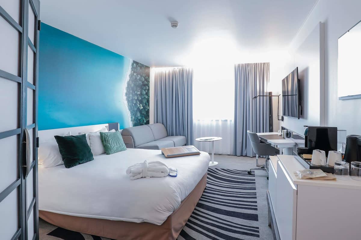 A hotel room with a bed, a desk, a chair, a small table, a TV, and a window with curtains.