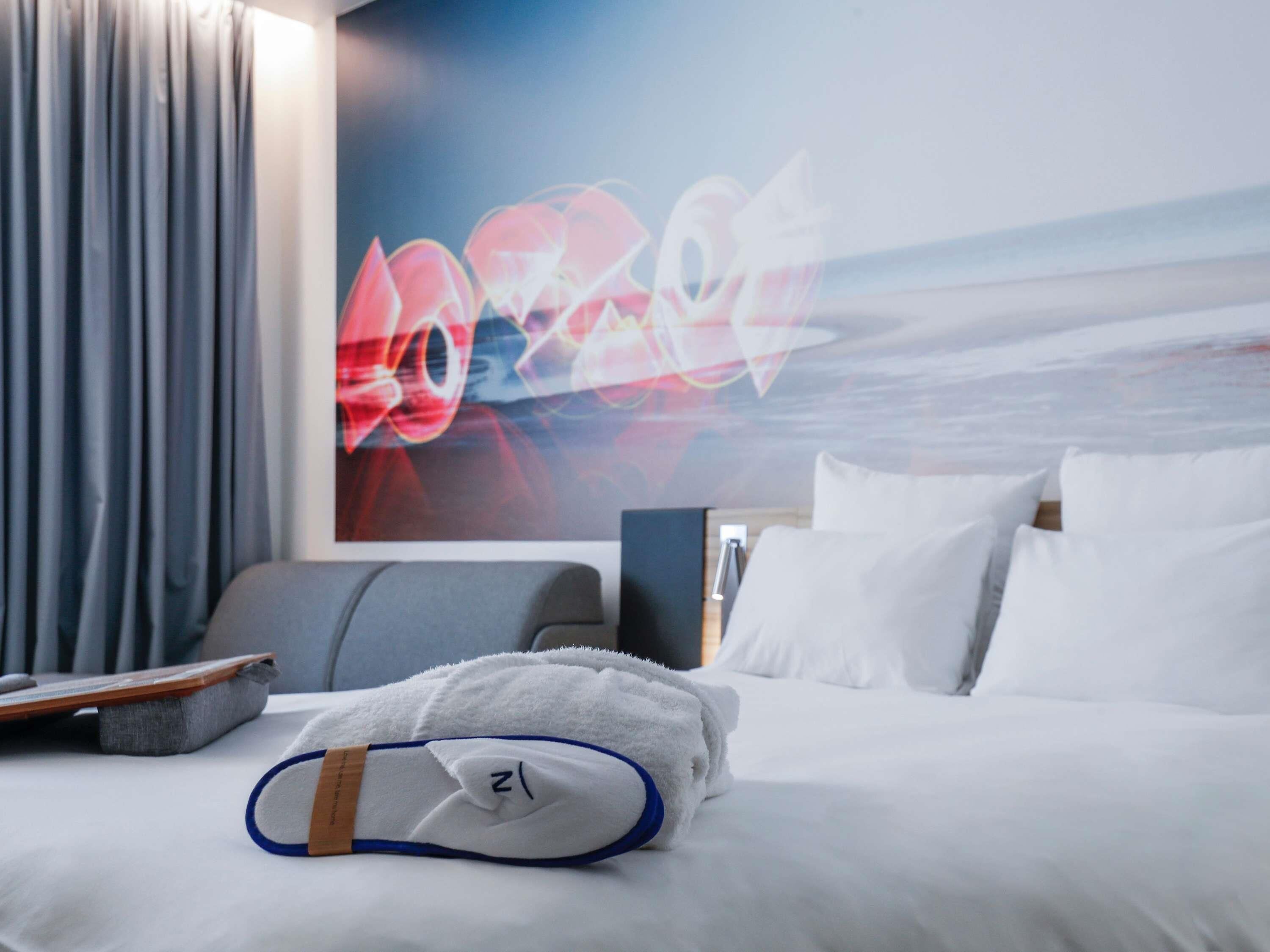 A hotel room with a bed, a towel, and a pair of slippers.