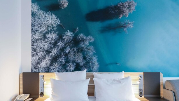 Una habitación de hotel con una cama, dos almohadas blancas, un cabecero de madera, una mesita de noche con un teléfono y un mural en la pared con un bosque.